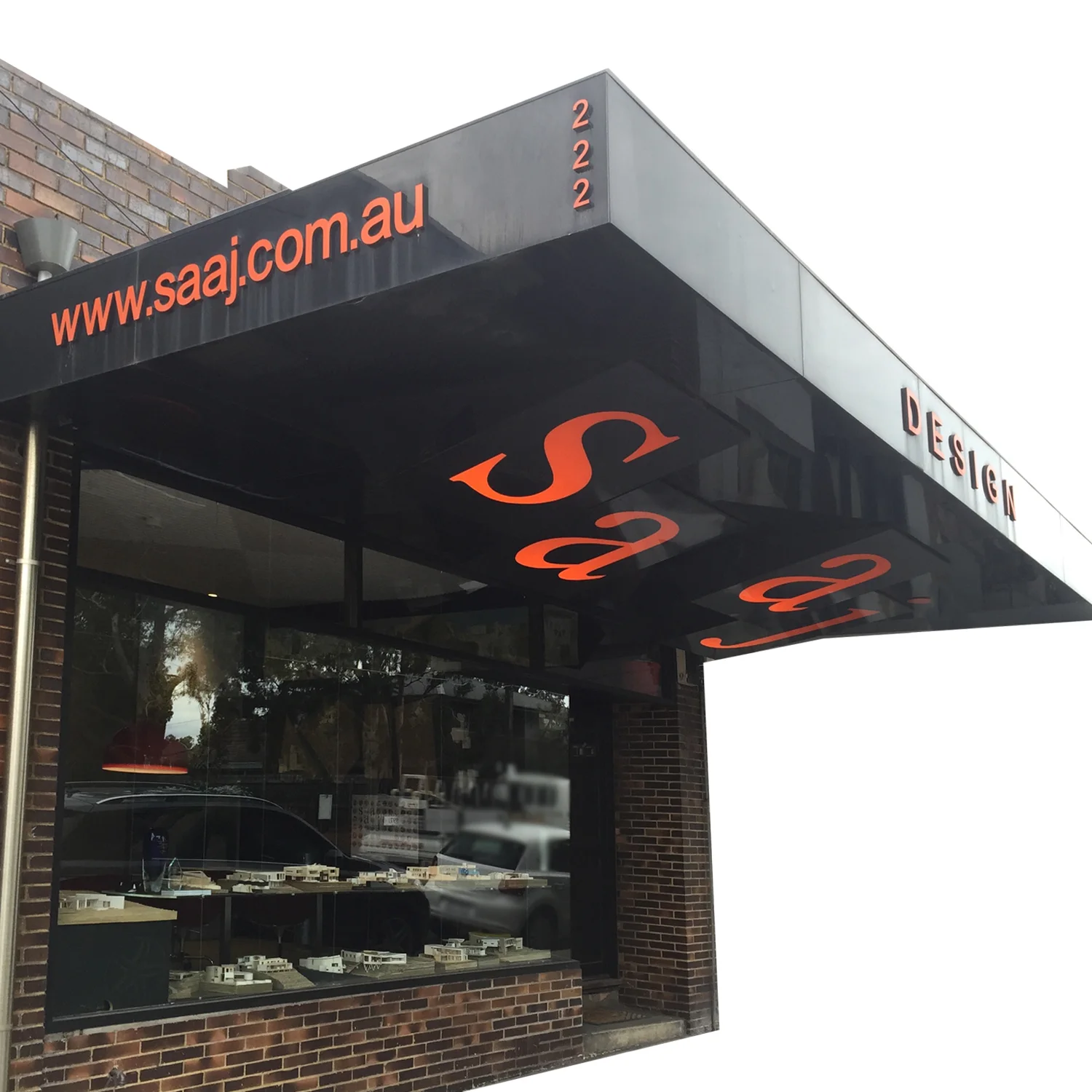 Storefront with brick exterior, large display window showing model buildings inside, black awning with orange text 'www.saj.com.au' and red 2222, and the partially visible word 'DESIGN' on the right side.