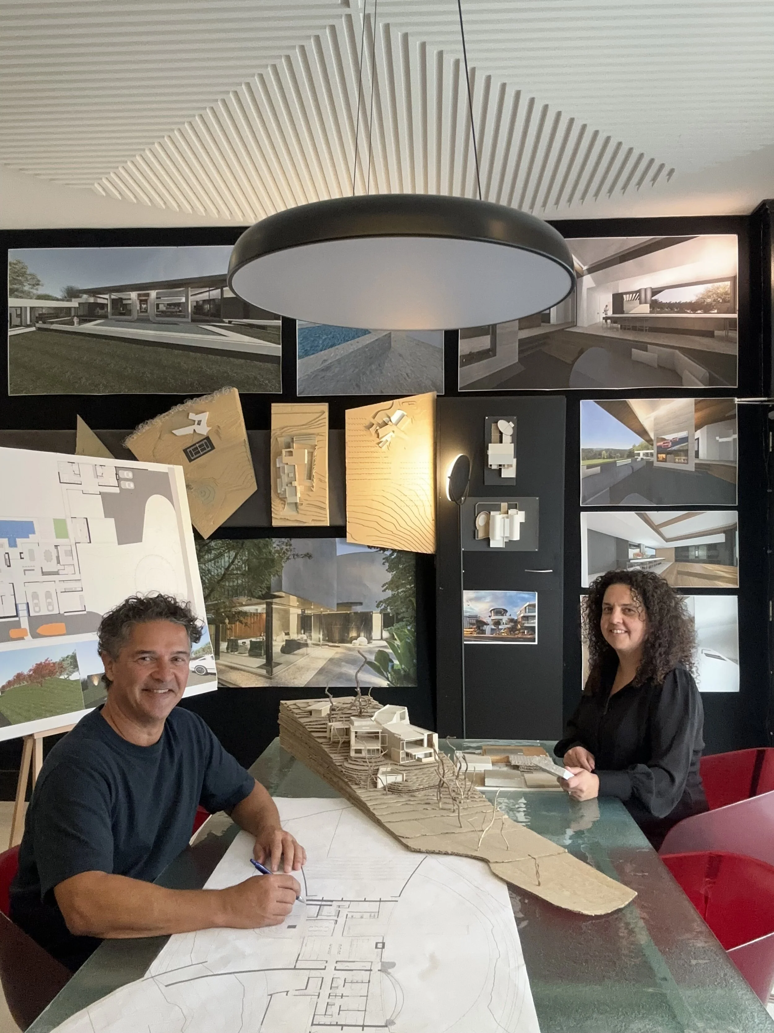 Two people sitting at a table with an architectural scale model and blueprints, with architectural renderings and plans displayed on a black wall behind them.