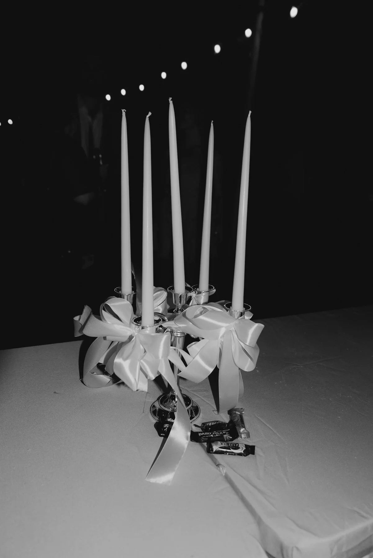 A black and white photo of a table with a decorative candelabrum holding five tall, unlit candles adorned with large satin bows and ribbons, with candy bars placed at the base.