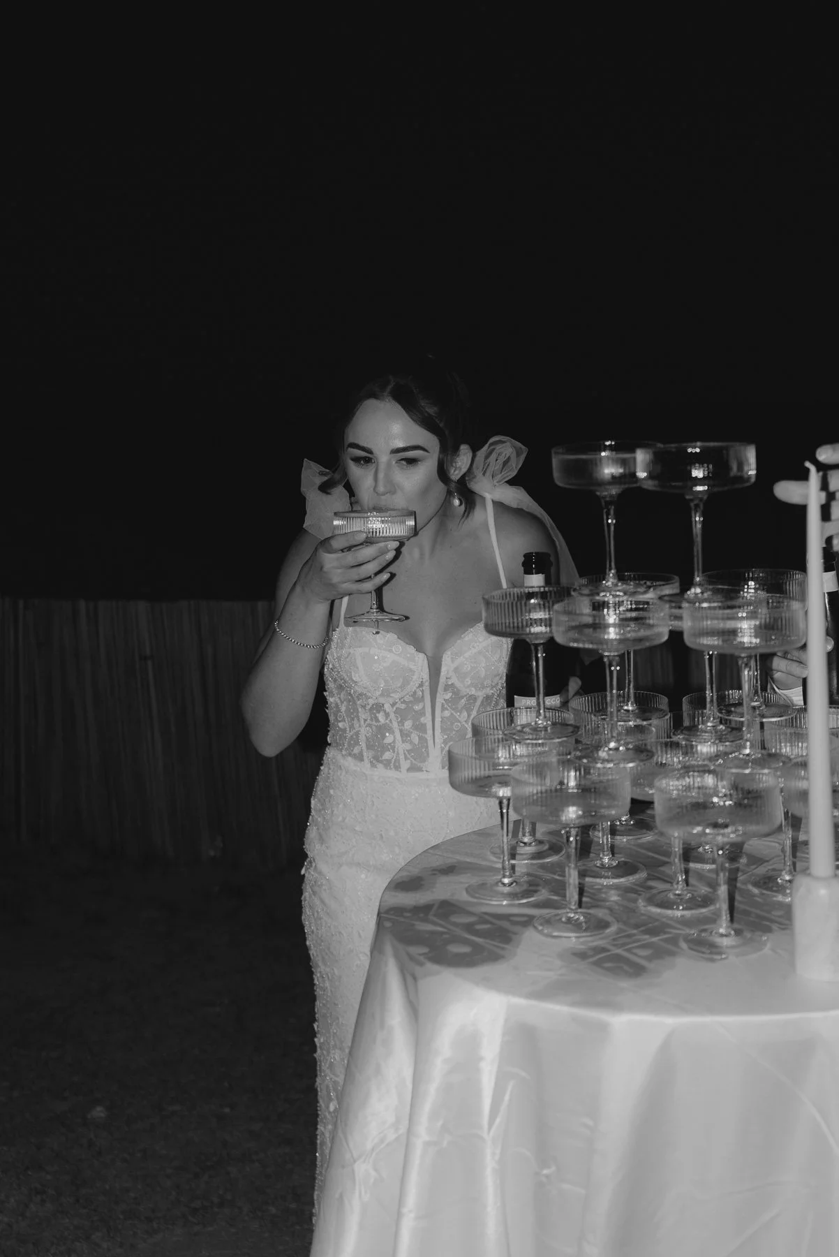 A woman in a white dress holds a glass and appears to be about to drink it, standing next to a table with stacked glasses and a white candle. The photo is in black and white.