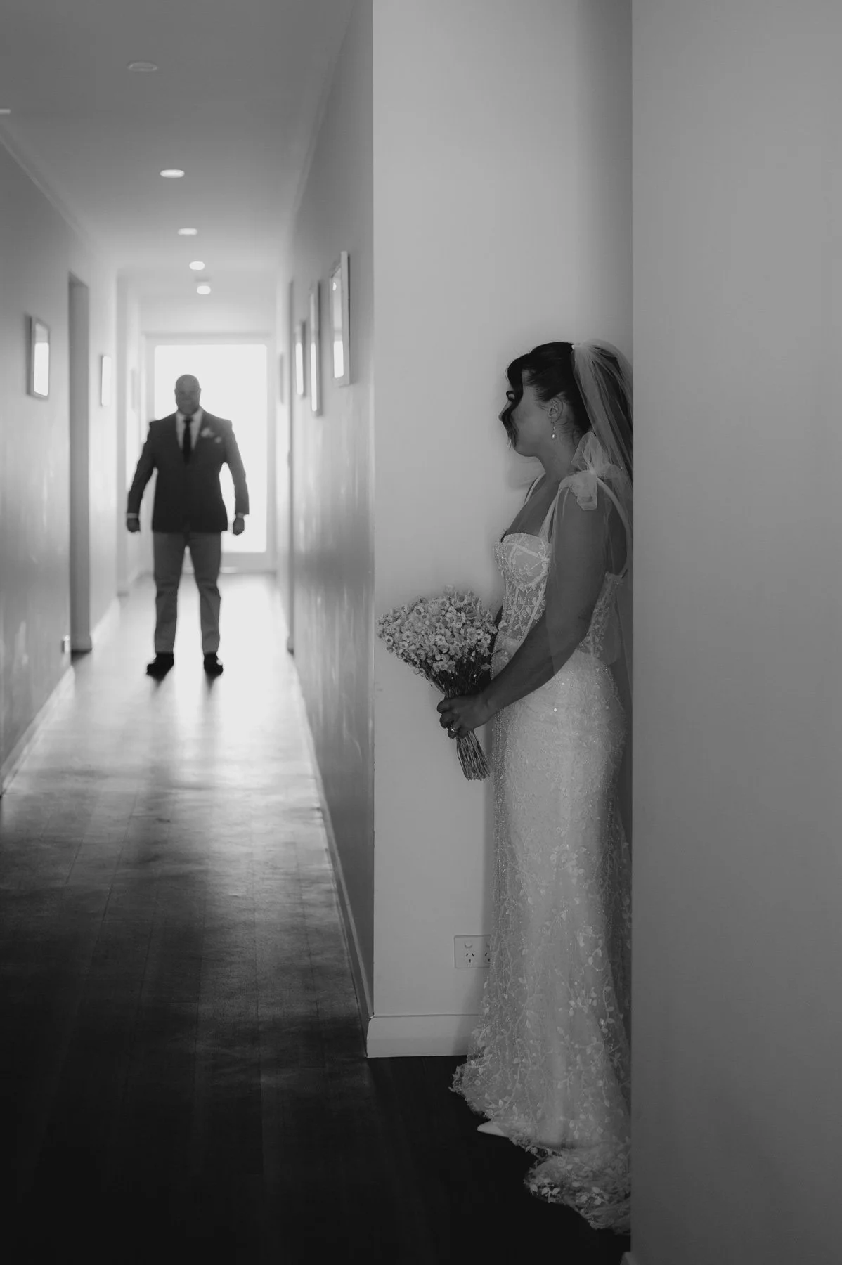 Black and white photo of a bride in wedding dress standing by a wall, holding a bouquet, looking at a man approaching from the end of a hallway.