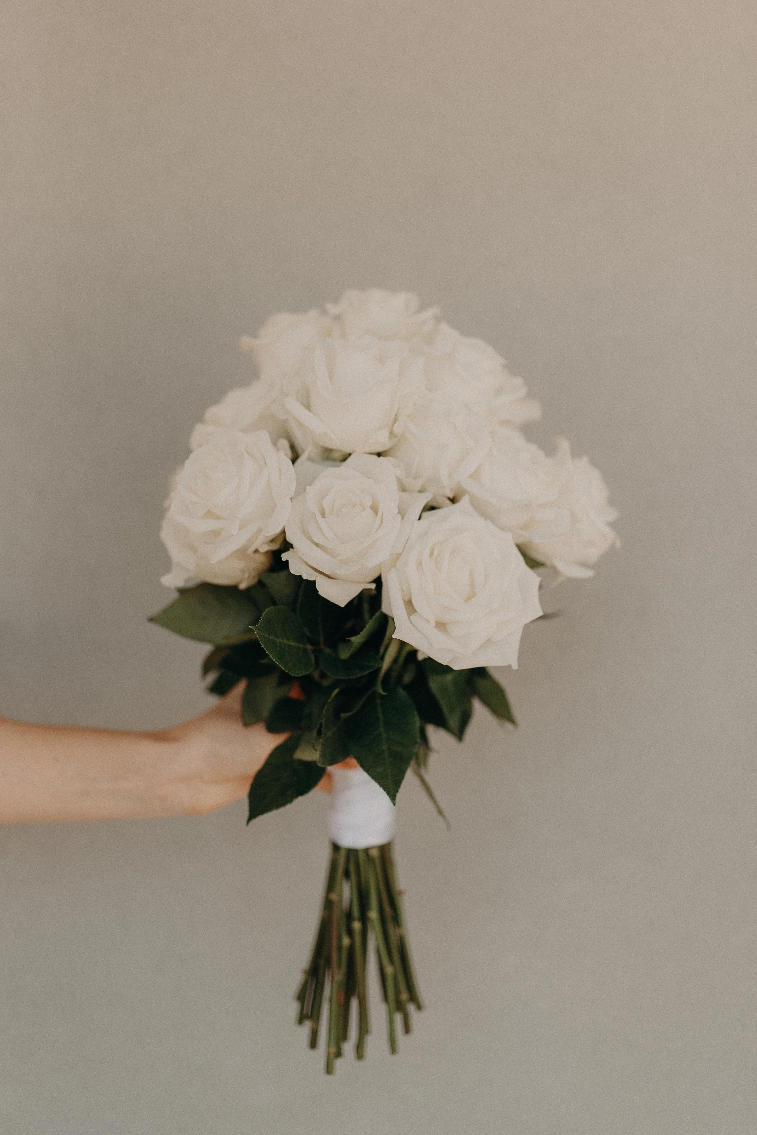 Hand holding a bouquet of white roses with green leaves, wrapped with white ribbon.