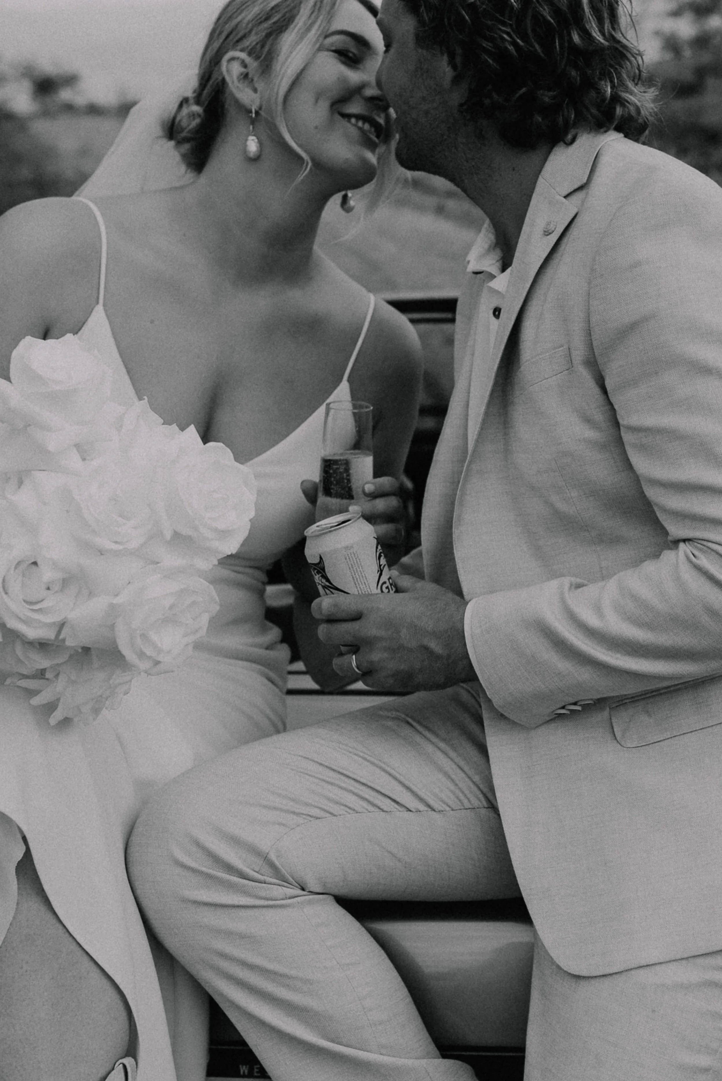Black and white photo of a bride and groom close together, smiling, with the bride holding a bouquet of roses and a glass, and the groom holding a can and a drink glass. The bride is wearing a dress with thin straps and earrings, and the groom is in 