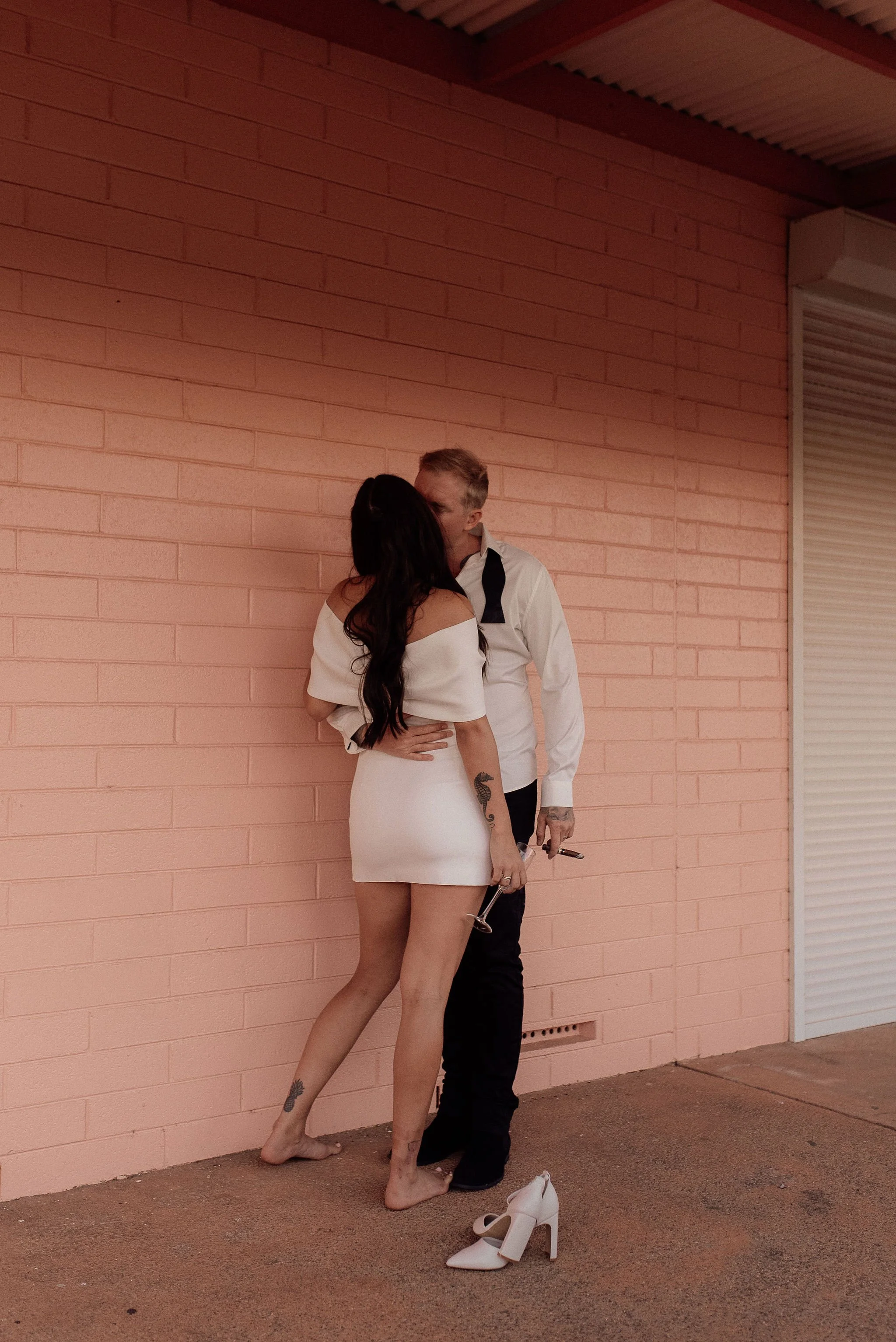 A couple kissing in front of a pink brick wall. The woman is in a white mini dress and is barefoot, holding white high heels. The man is in a white shirt with a black tie and black pants, holding a glass of wine.