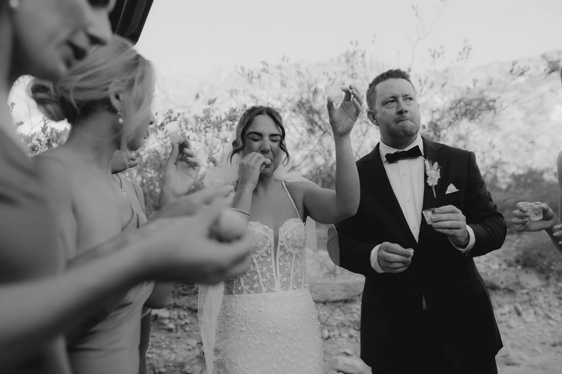 Black and white photo of a wedding reception outdoors, with women and a man in formal attire, some holding drinks, some eating, one woman in a wedding dress
