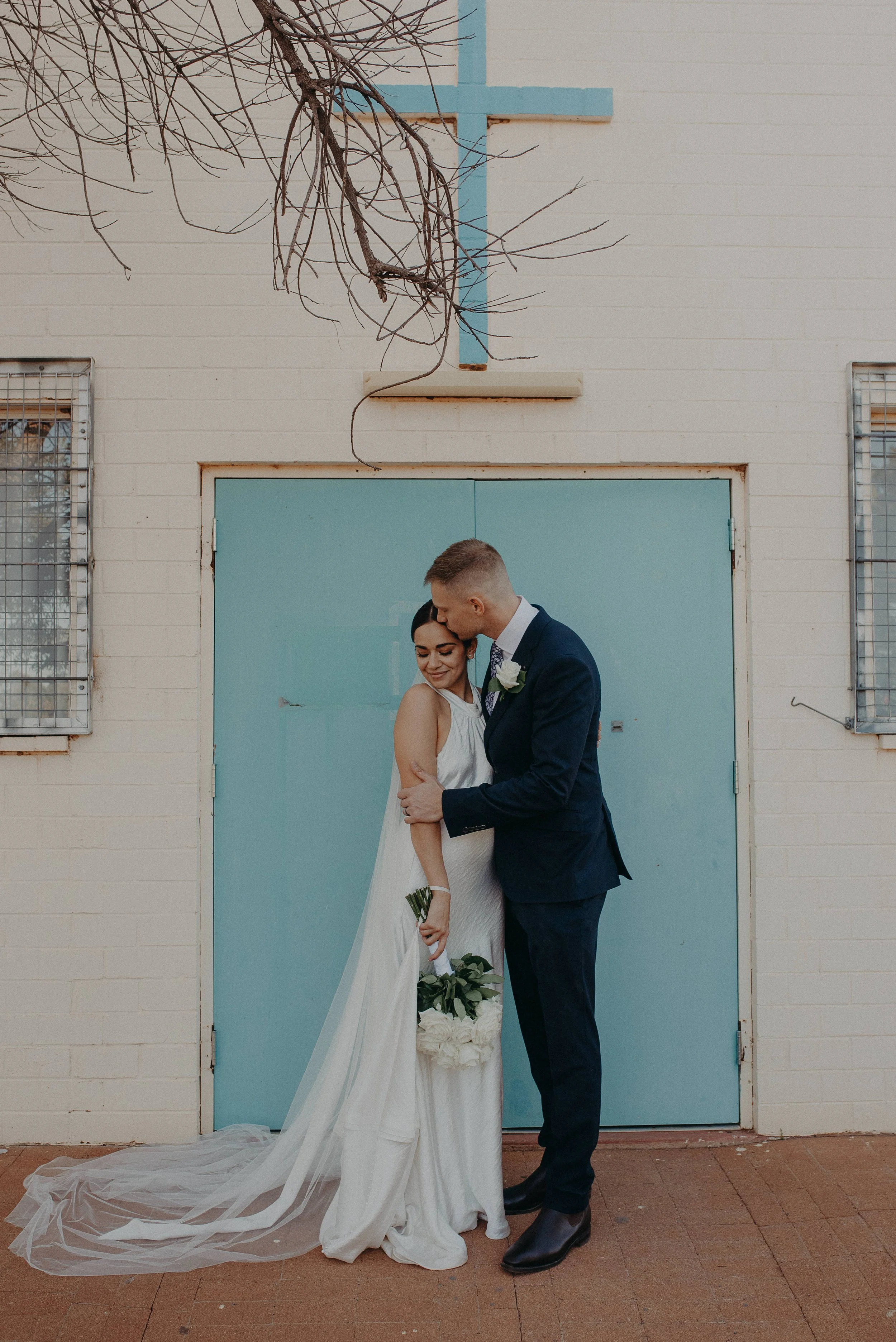 A bride and groom standing close together in front of a turquoise door, with the groom kissing the bride's forehead. The bride is holding a bouquet of white flowers and wearing a white gown with a long, flowing train. The groom is dressed in a dark b