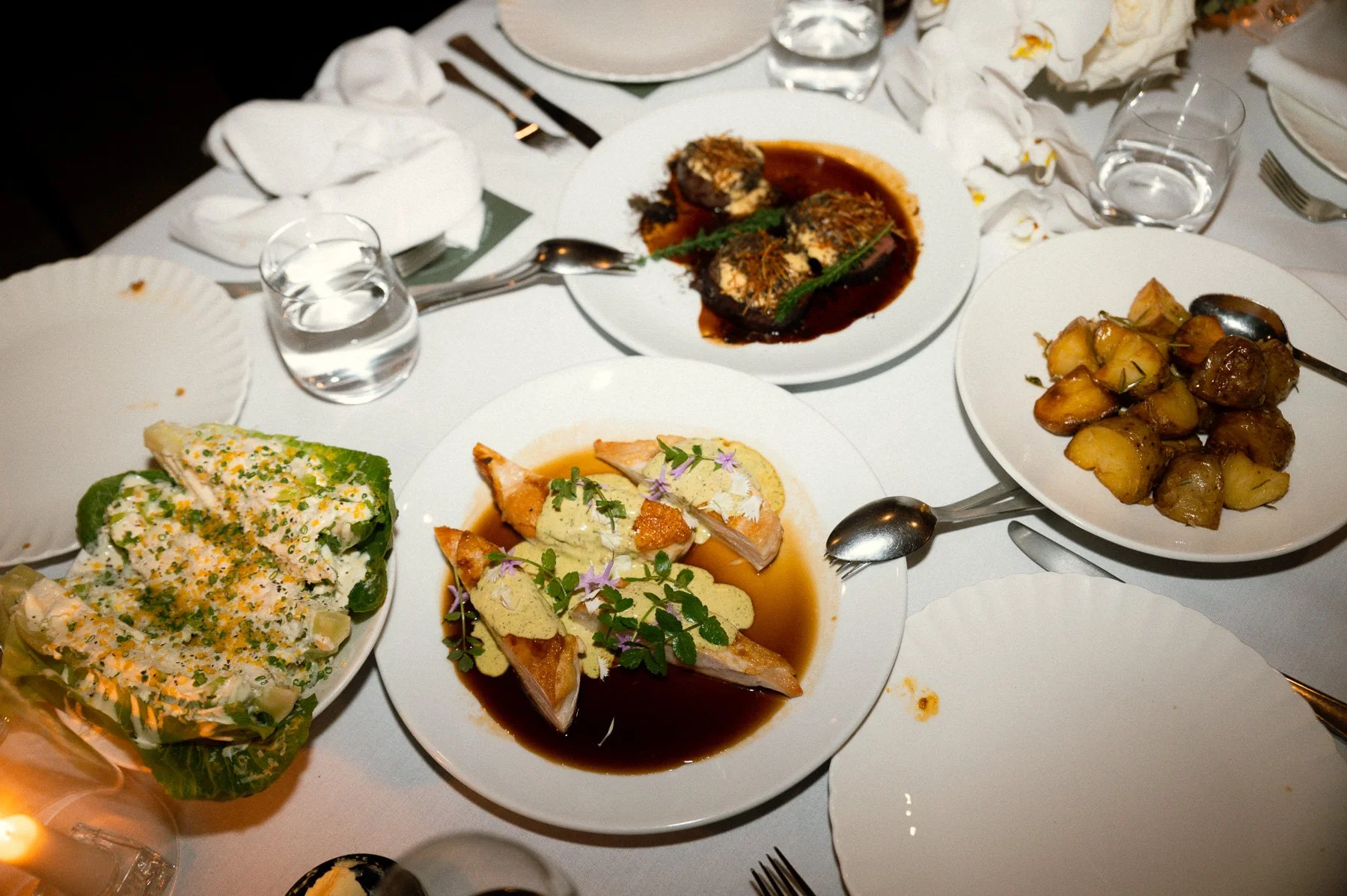 A dinner table with various plated dishes including a salad, chicken with sauce, roasted potatoes, a meat dish in sauce, and a vegetable dish, set with utensils, glasses of water, and napkins.