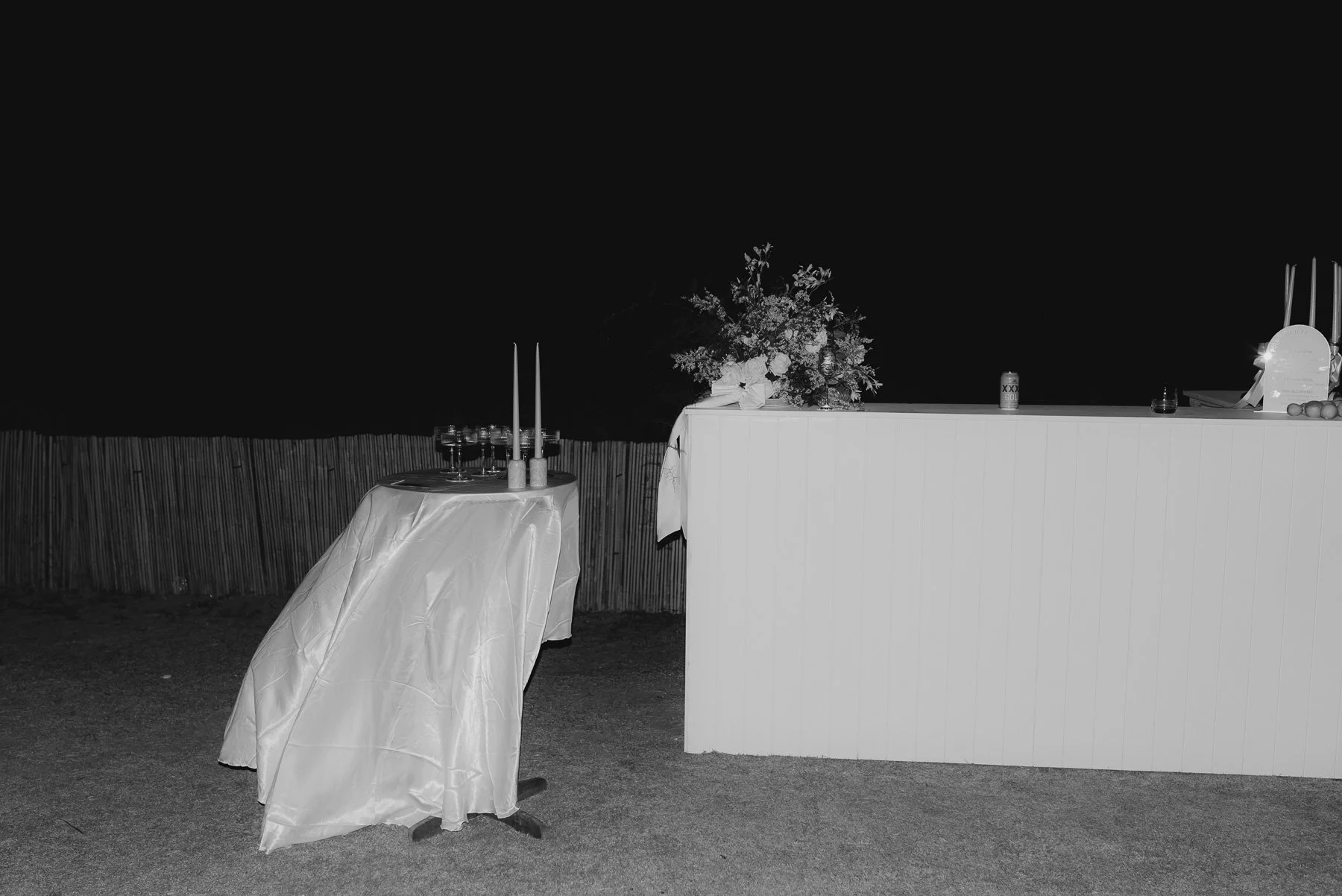 Nighttime outdoor bar setup with a lit flower arrangement on a white counter and a small table with glasses and candles.