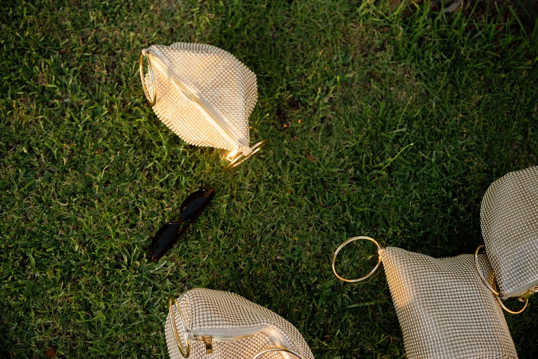 Three small gold handbags and a pair of sunglasses on green grass, with one handbag having a circular gold handle.