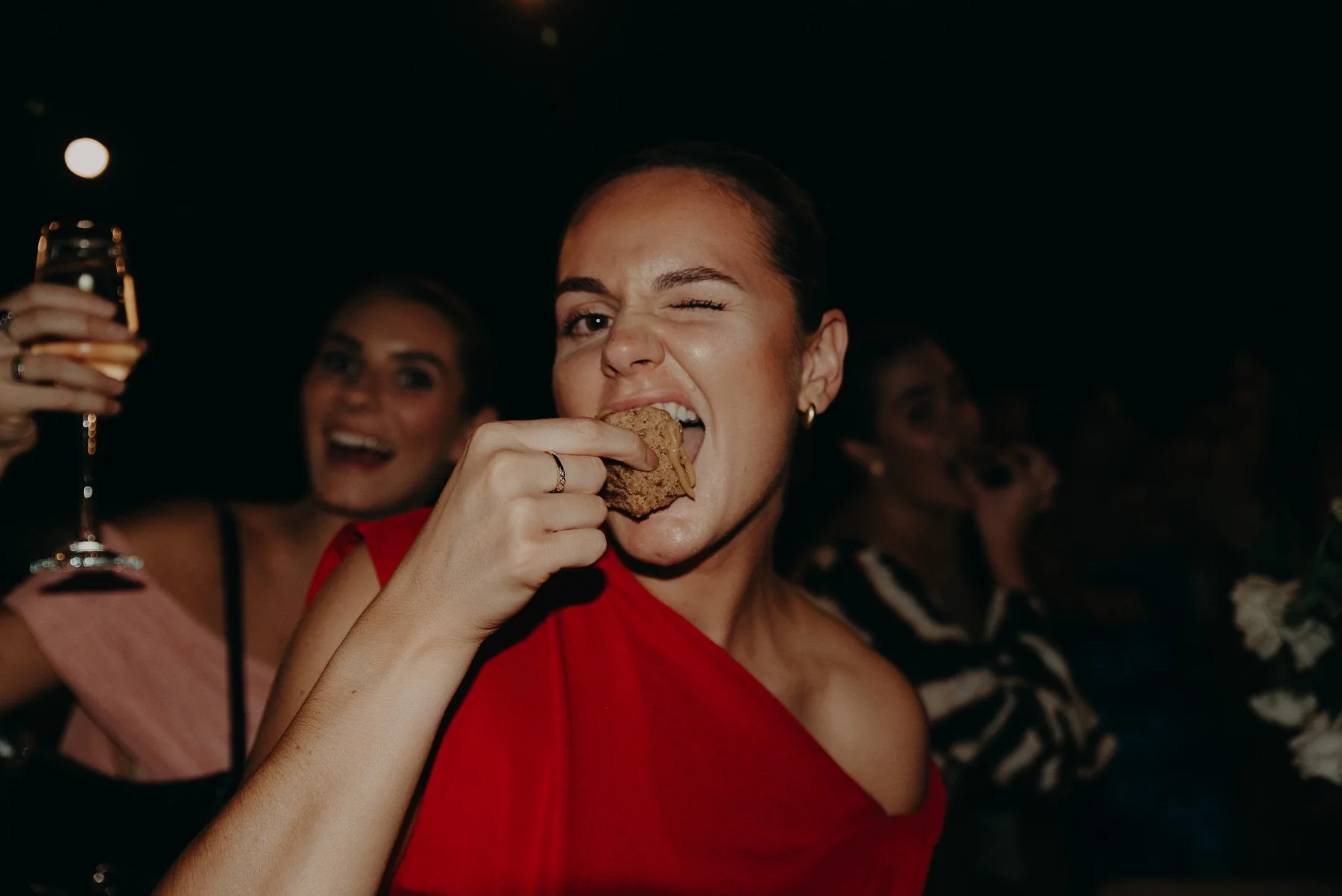 Woman in a red dress eating a cookie at a party, with two women in the background, one holding a glass of champagne and smiling.