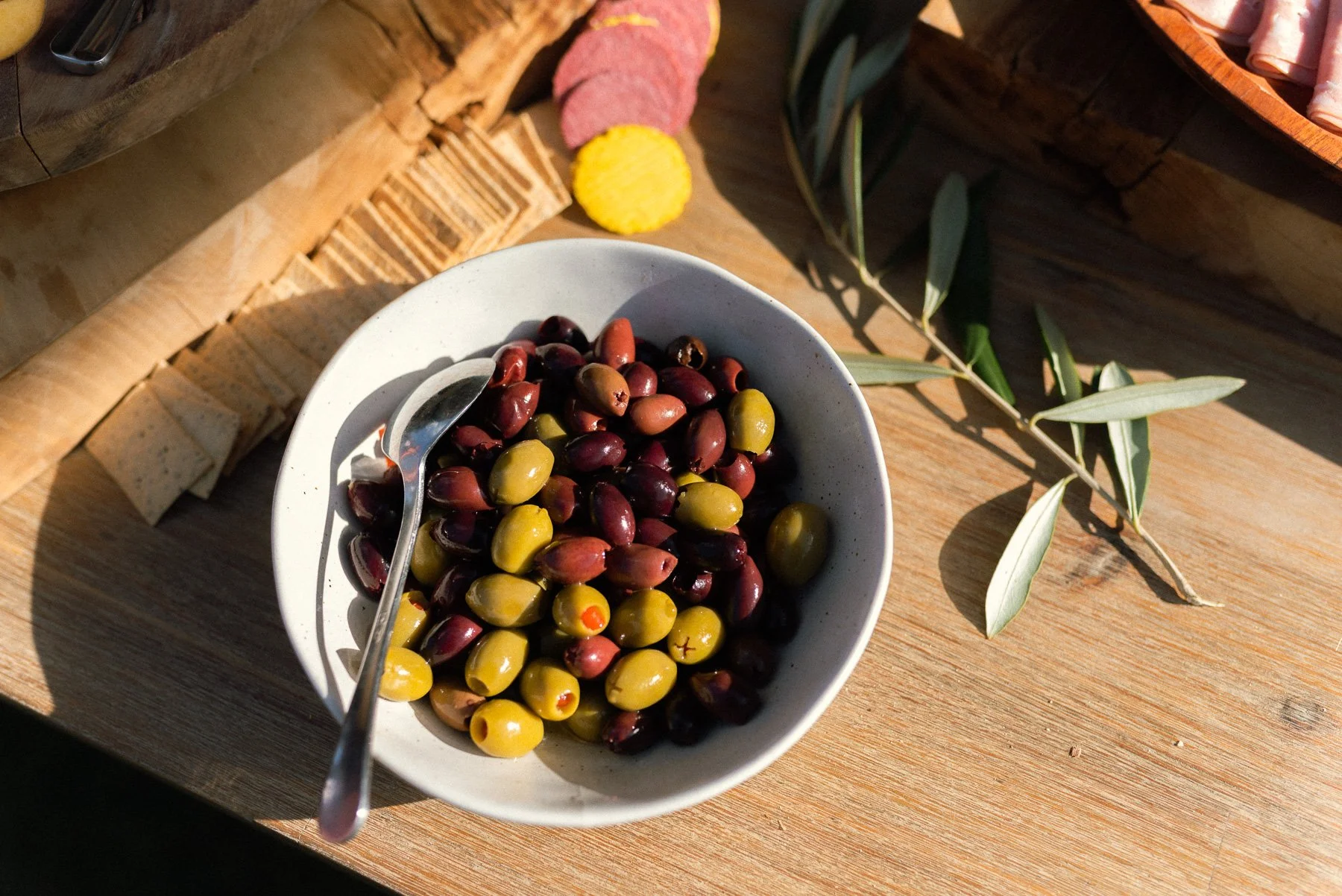 A bowl of mixed green and black olives with a silver spoon inside, placed on a wooden surface with sunlight and a branch with green leaves nearby.