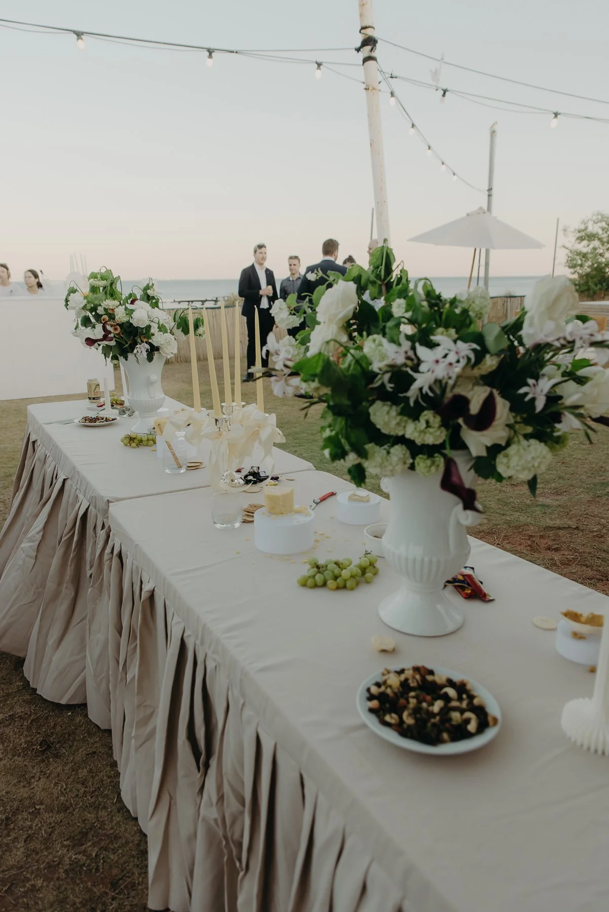 Long reception table with white tablecloth and large floral arrangements in white vases, set outdoors near a beach with string lights overhead and people in formal attire in the background.