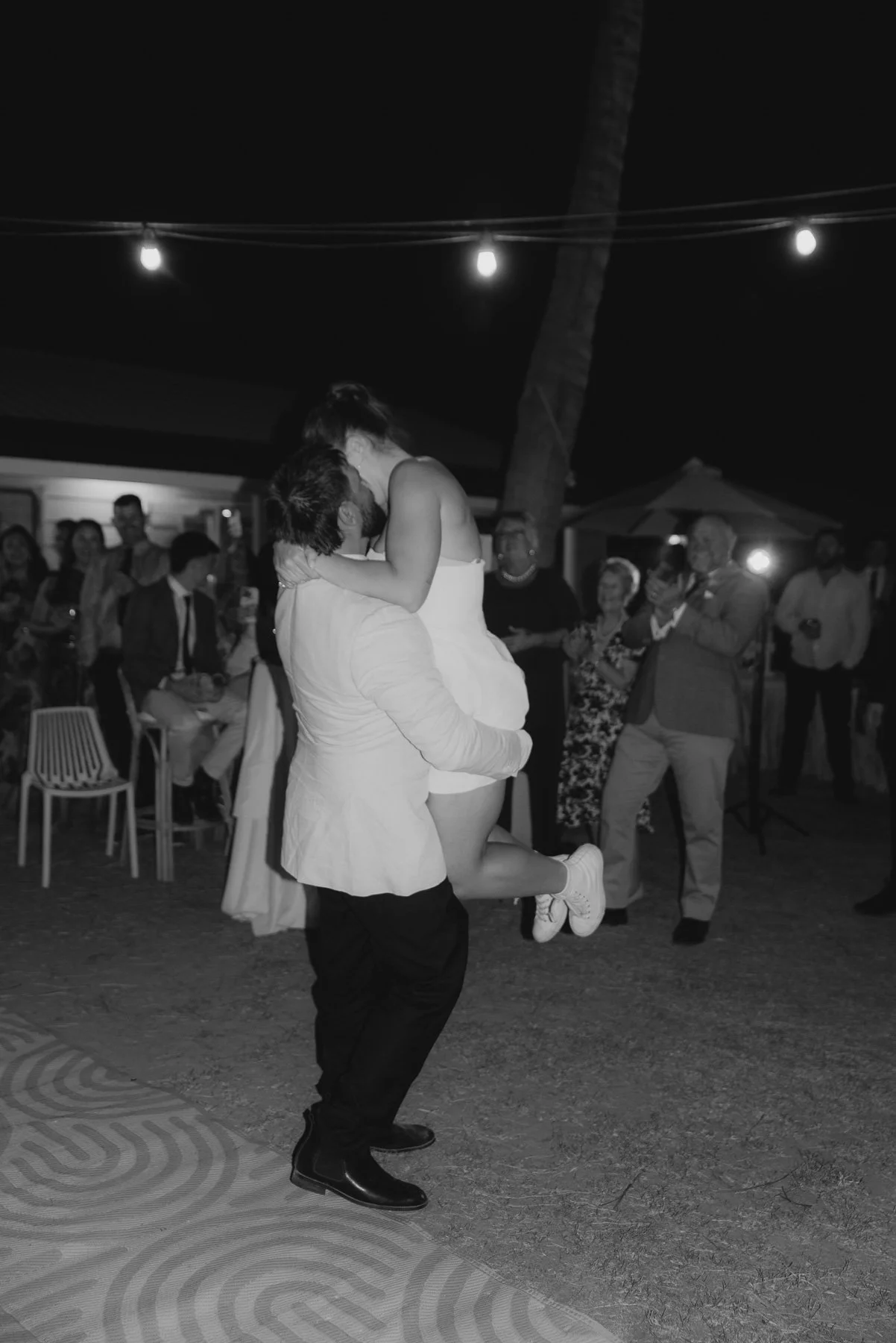 A man is lifting a woman at an outdoor wedding reception at night, surrounded by guests clapping and smiling. String lights hang overhead.