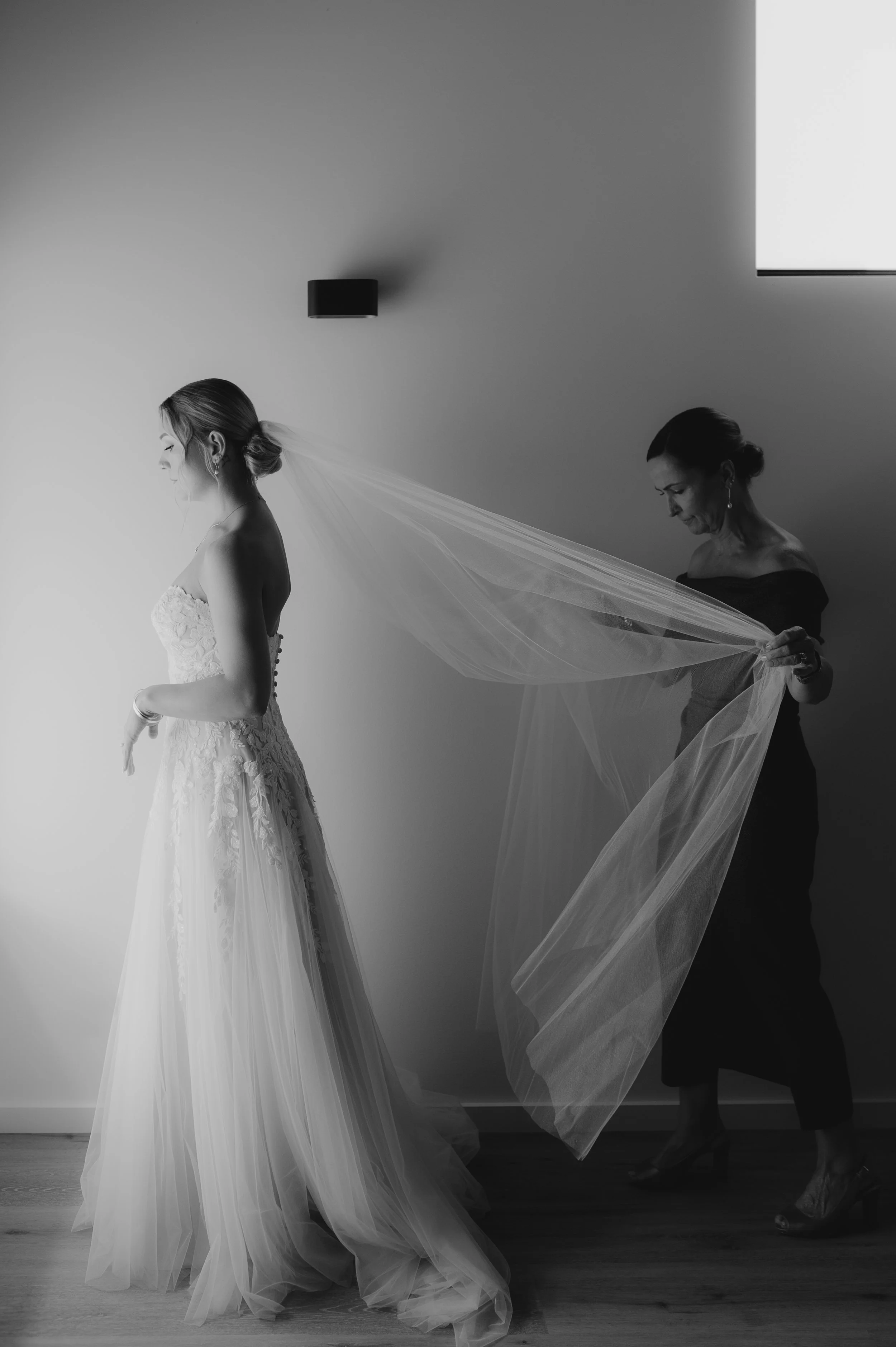 A bride in a wedding dress standing sideways while a woman behind her helps her with her veil, in a minimalistic room with a wall and wooden floor.