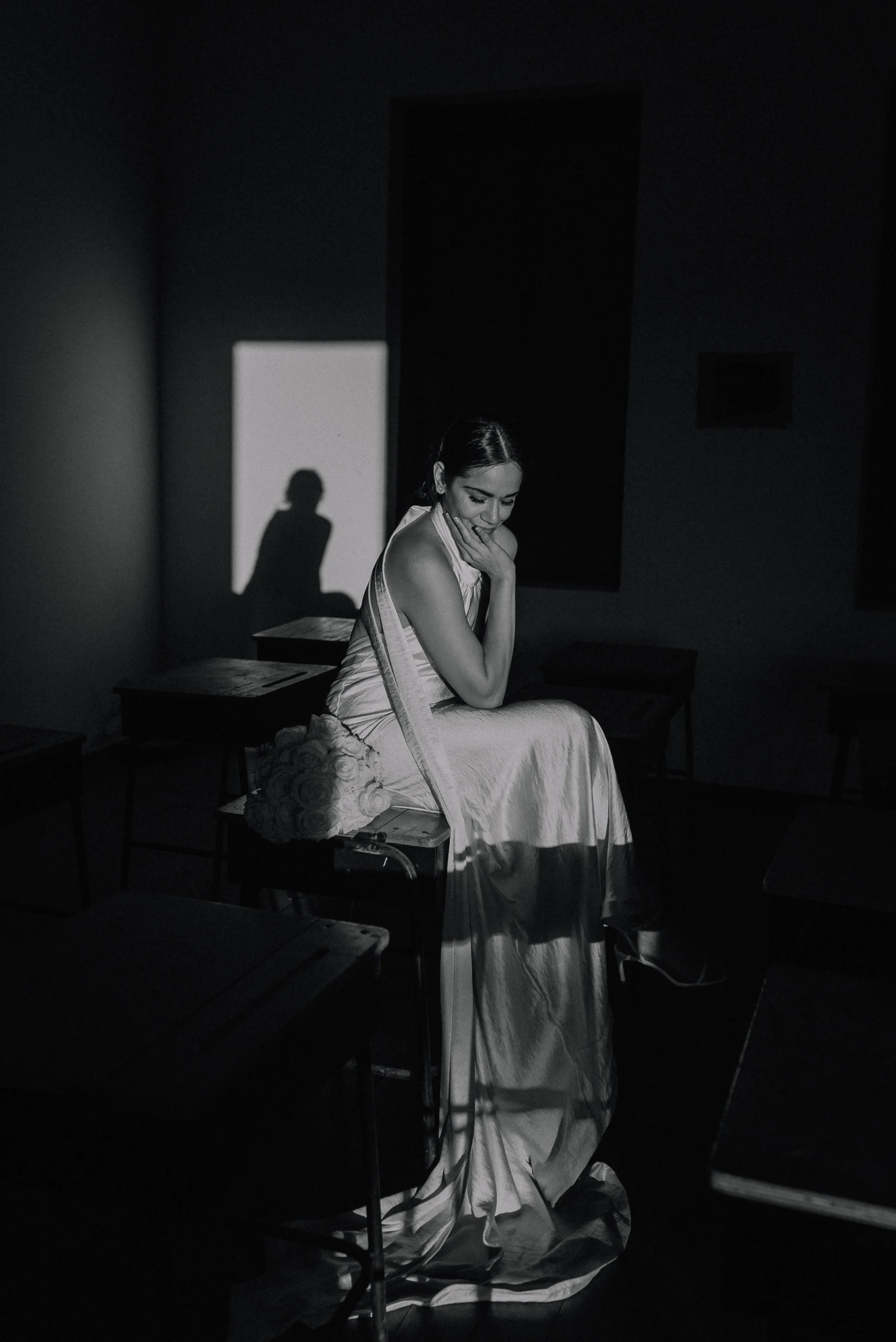 A woman in a light-colored dress sitting on a table in a dark room, with a bouquet of roses beside her, illuminated by a single light source creating shadows on the wall.