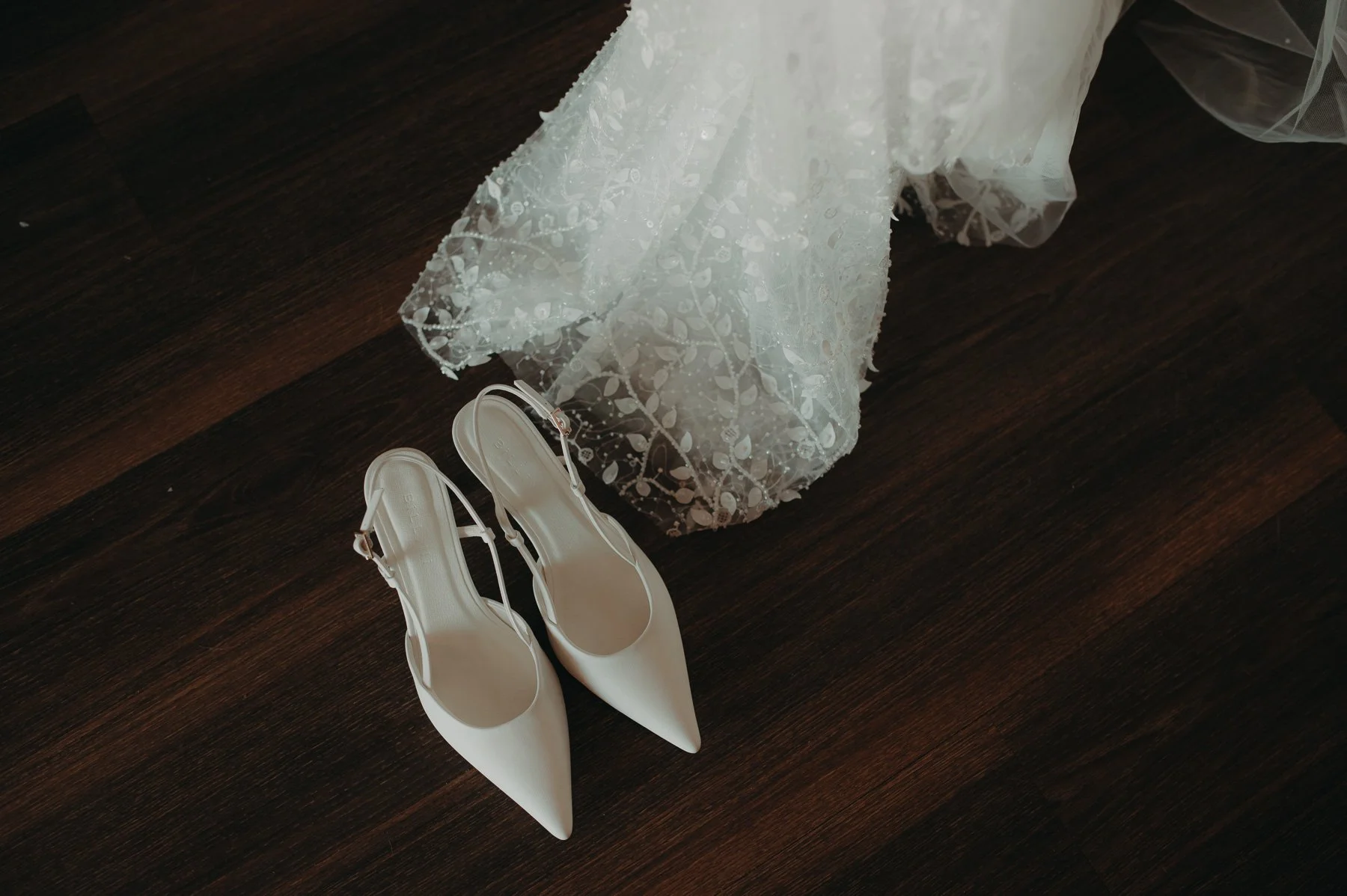White wedding shoes and a lace wedding dress on a dark wooden floor.