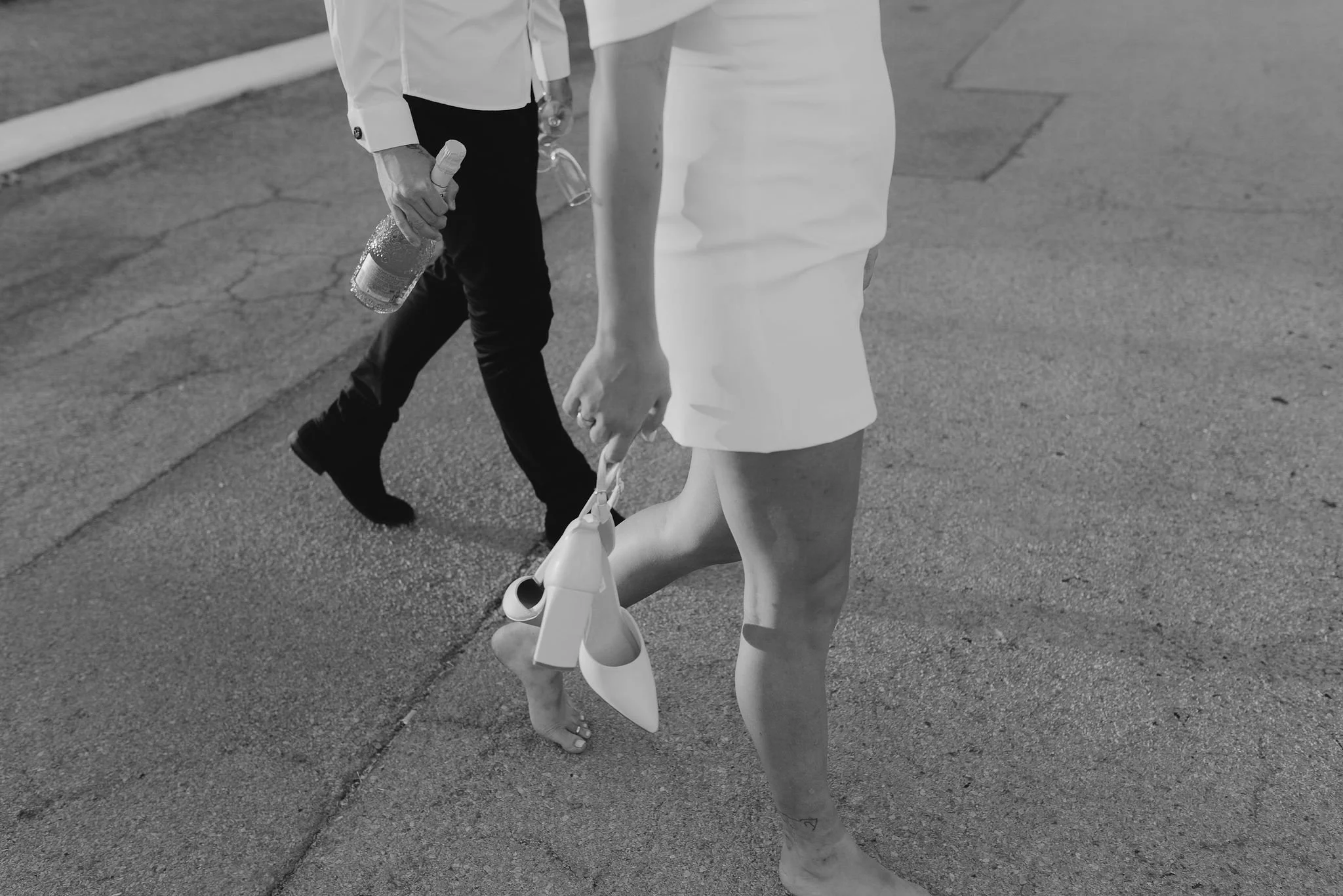 Black and white photo of a person in a white dress walking barefoot on the street, carrying a white bag and a rolled-up poster. Another person, dressed in dark pants and a light shirt, is walking nearby, holding a water bottle.