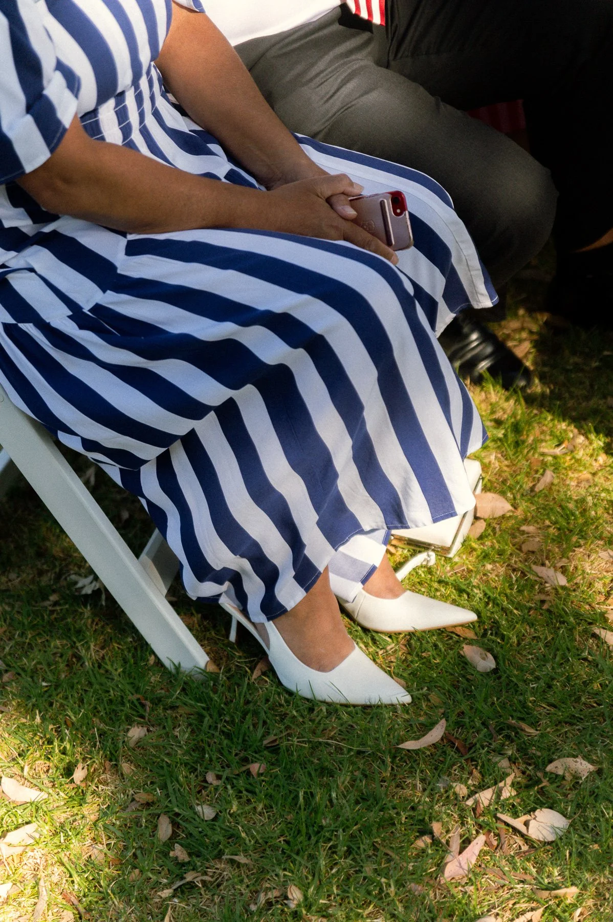 Woman wearing a blue and white striped dress and white high heels sitting on a white chair outdoors, holding a smartphone.