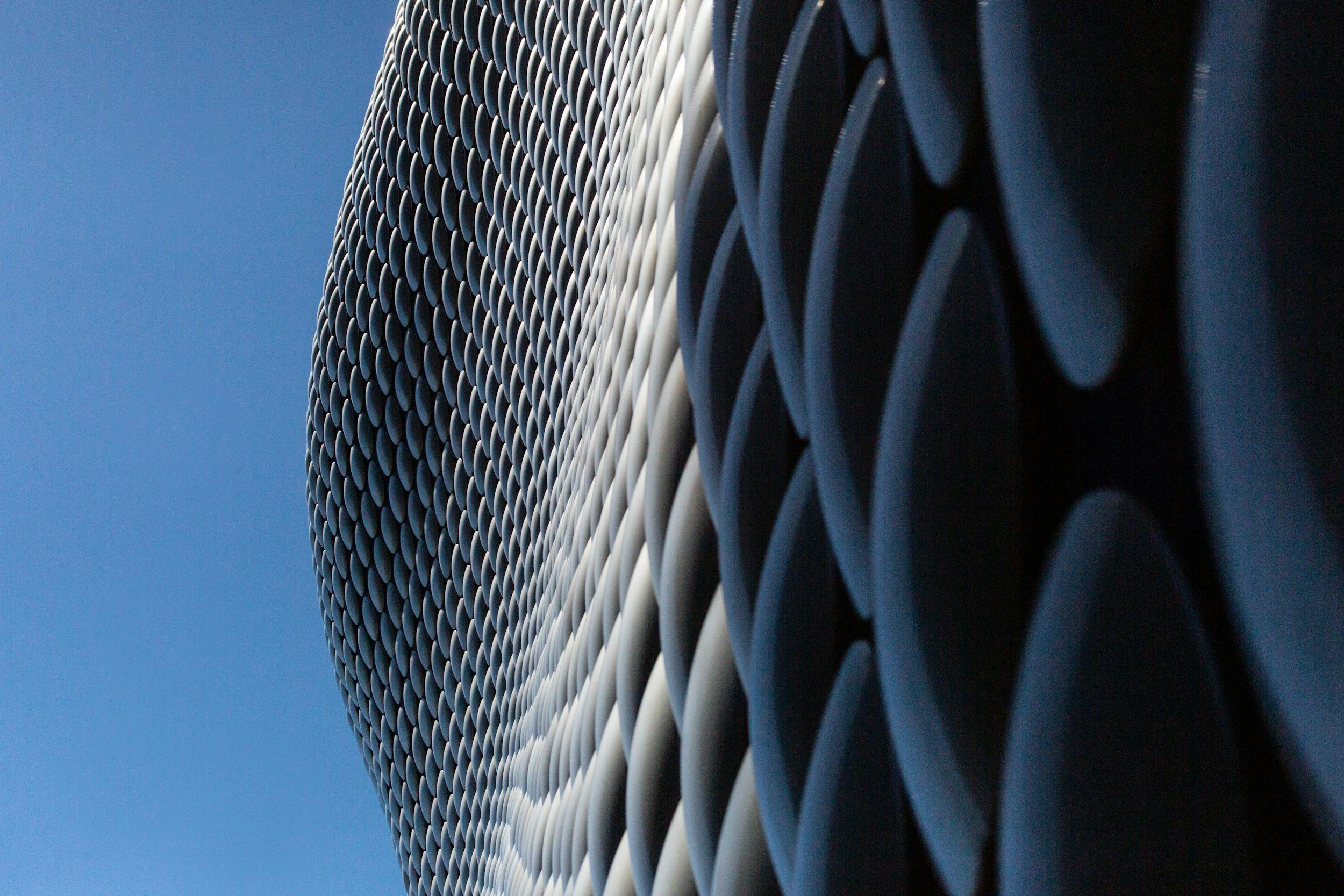 Close-up of a modern building's facade with a pattern of white, curved, overlapping panels against a clear blue sky.