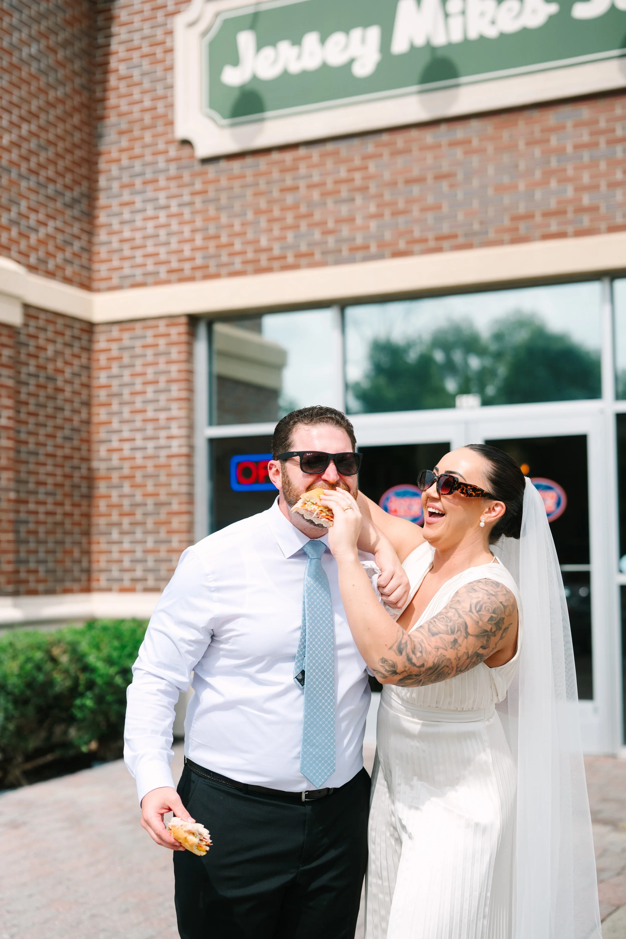 A couple in wedding attire, man in shirt and tie, woman in white dress with tattoos, laughing and sharing a sandwich outside Jersey Mike's Subs.