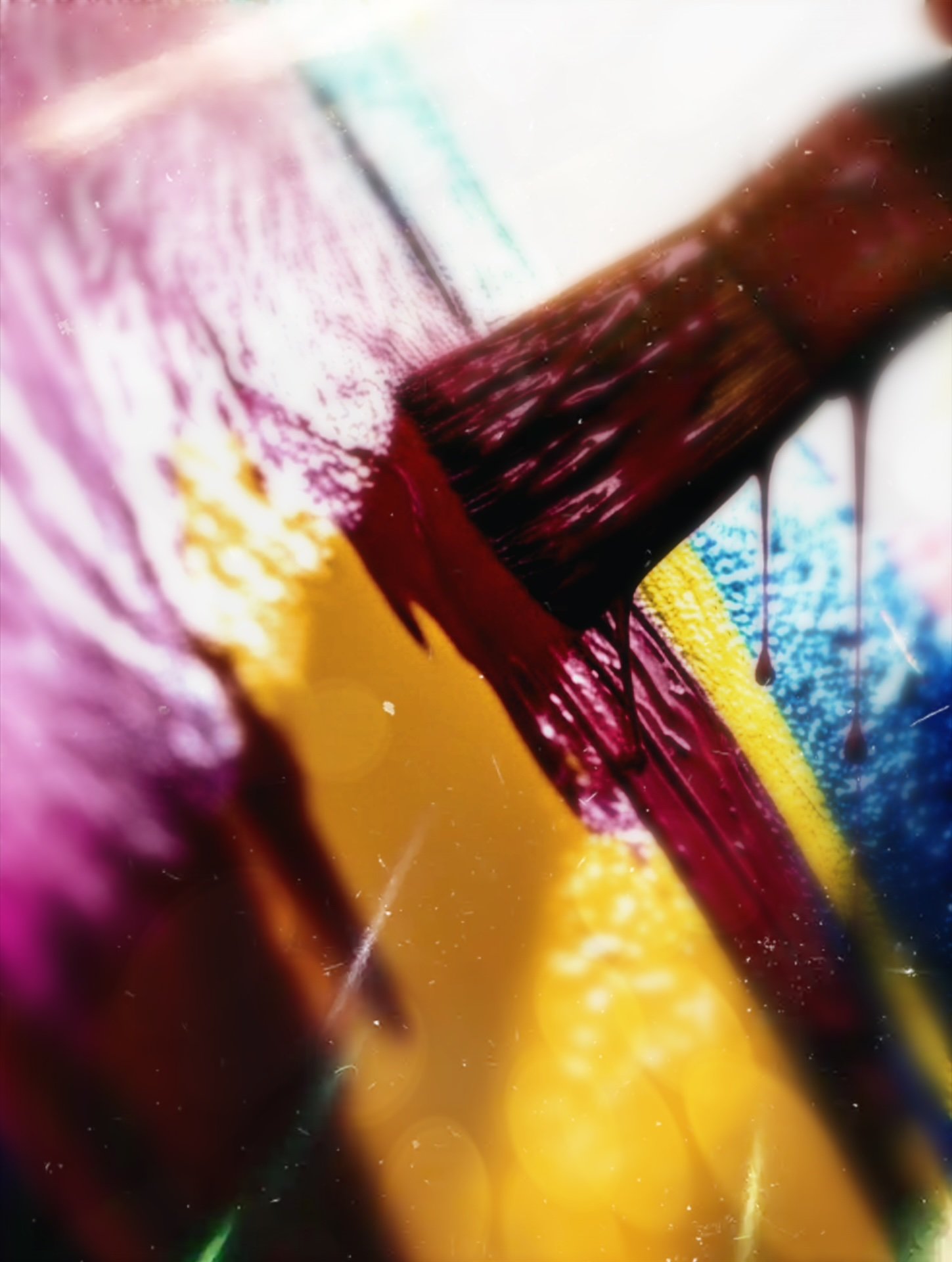 Close-up of a paintbrush applying dark red, purple, yellow, blue, and white paint with overlapping colors and textures.