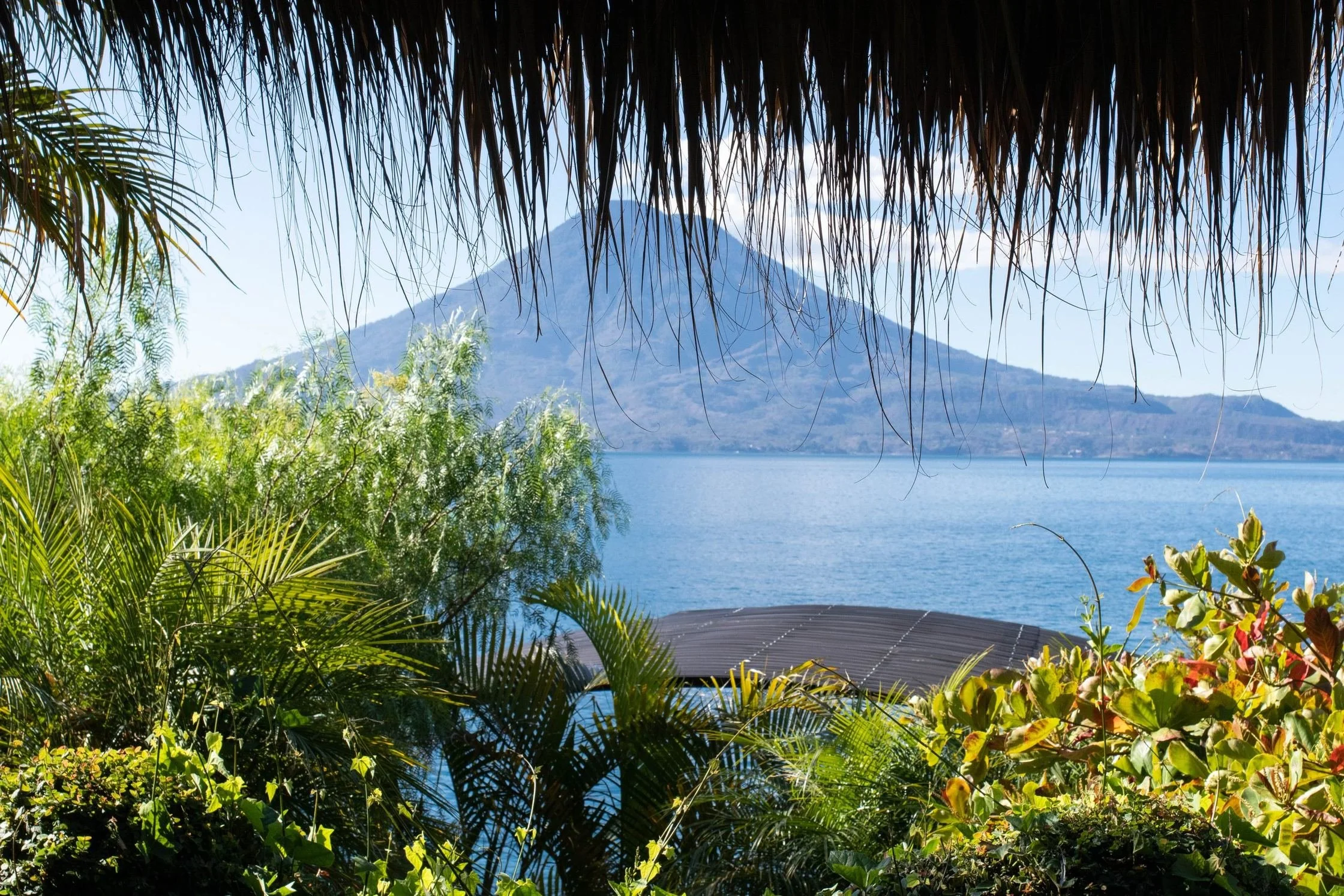 Volcano rising beyond a blue lake, framed by a thatched roof edge and lush tropical plants in the foreground