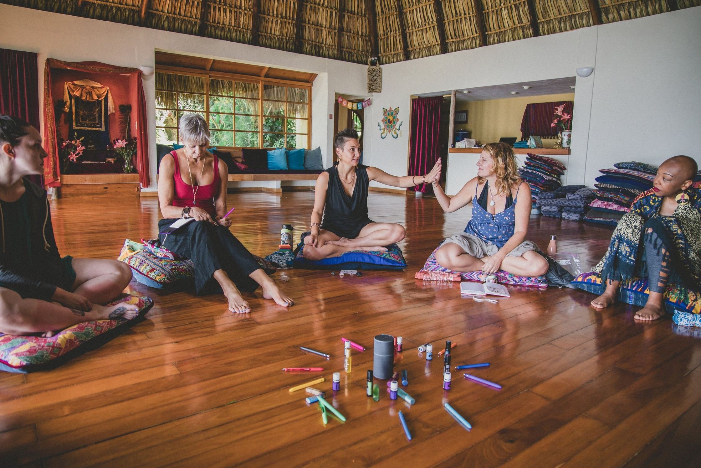 Five women sit on floor cushions in a circle in a wood-floored thatched-roof room, with markers and oil bottles in the center.