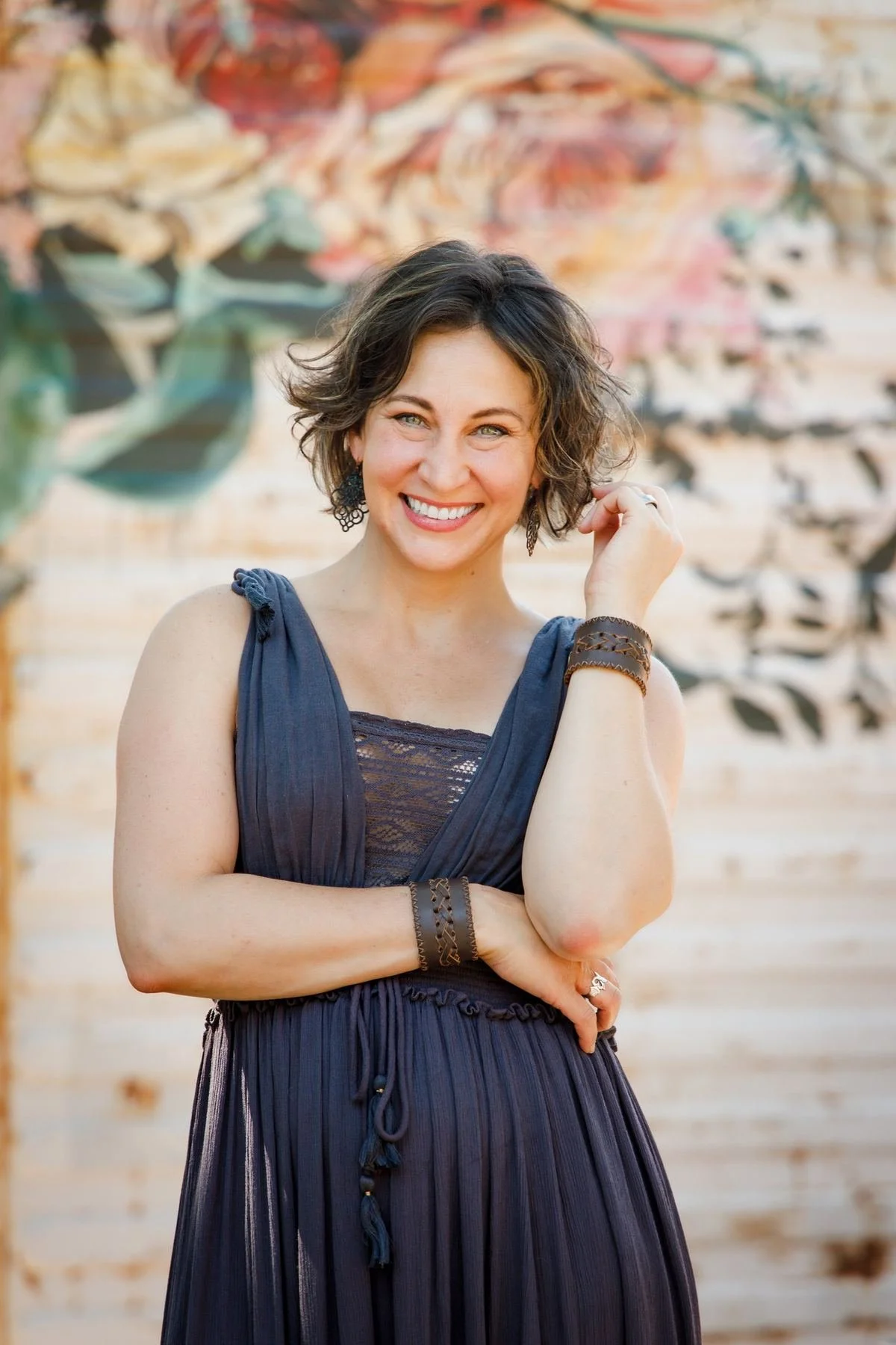 Smiling woman with short wavy brown hair, a gray-blue dress and leather cuffs, standing before a floral mural