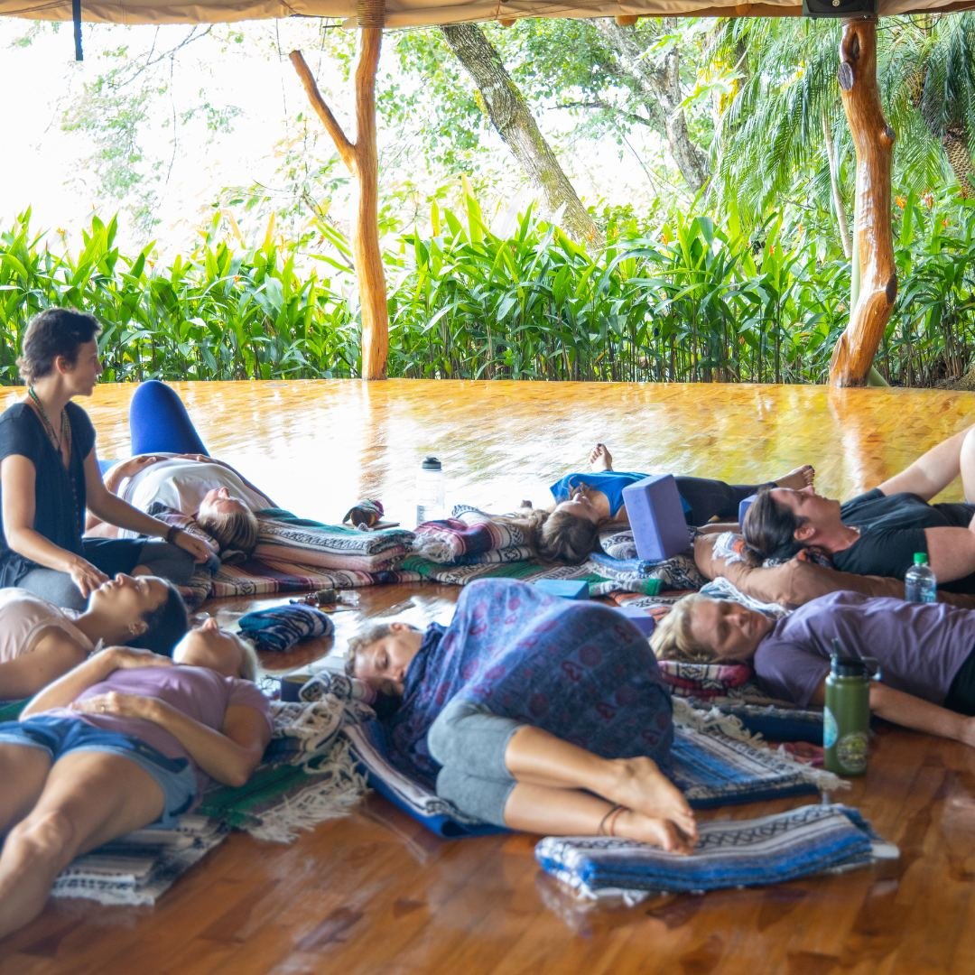 Ashlie Woods sits beside people resting on blankets on a wooden open air platform surrounded by tropical plants