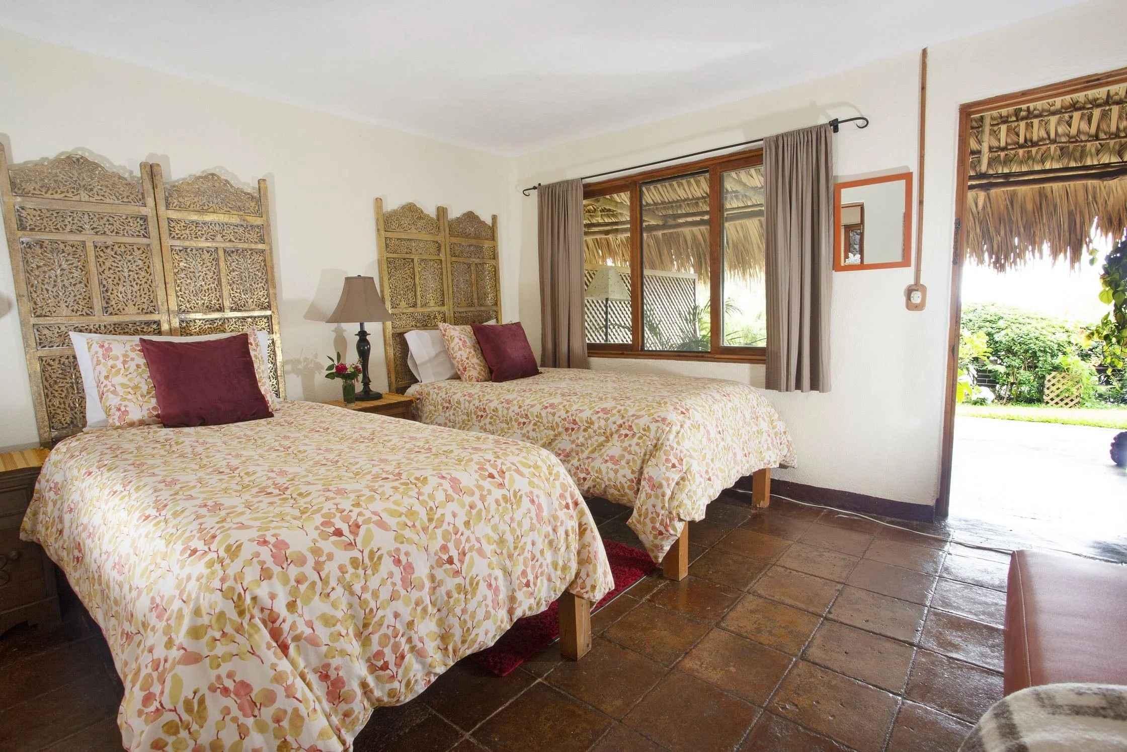 Two beds with floral bedspreads and carved wooden headboards in a tiled room with an open door to a garden