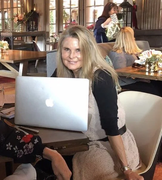 A woman with long blonde hair smiling at the camera, sitting at a table with a silver laptop in front of her. She is in a cozy, well-lit room with large windows and other people in the background, including a woman standing and another woman sitting with blonde hair.