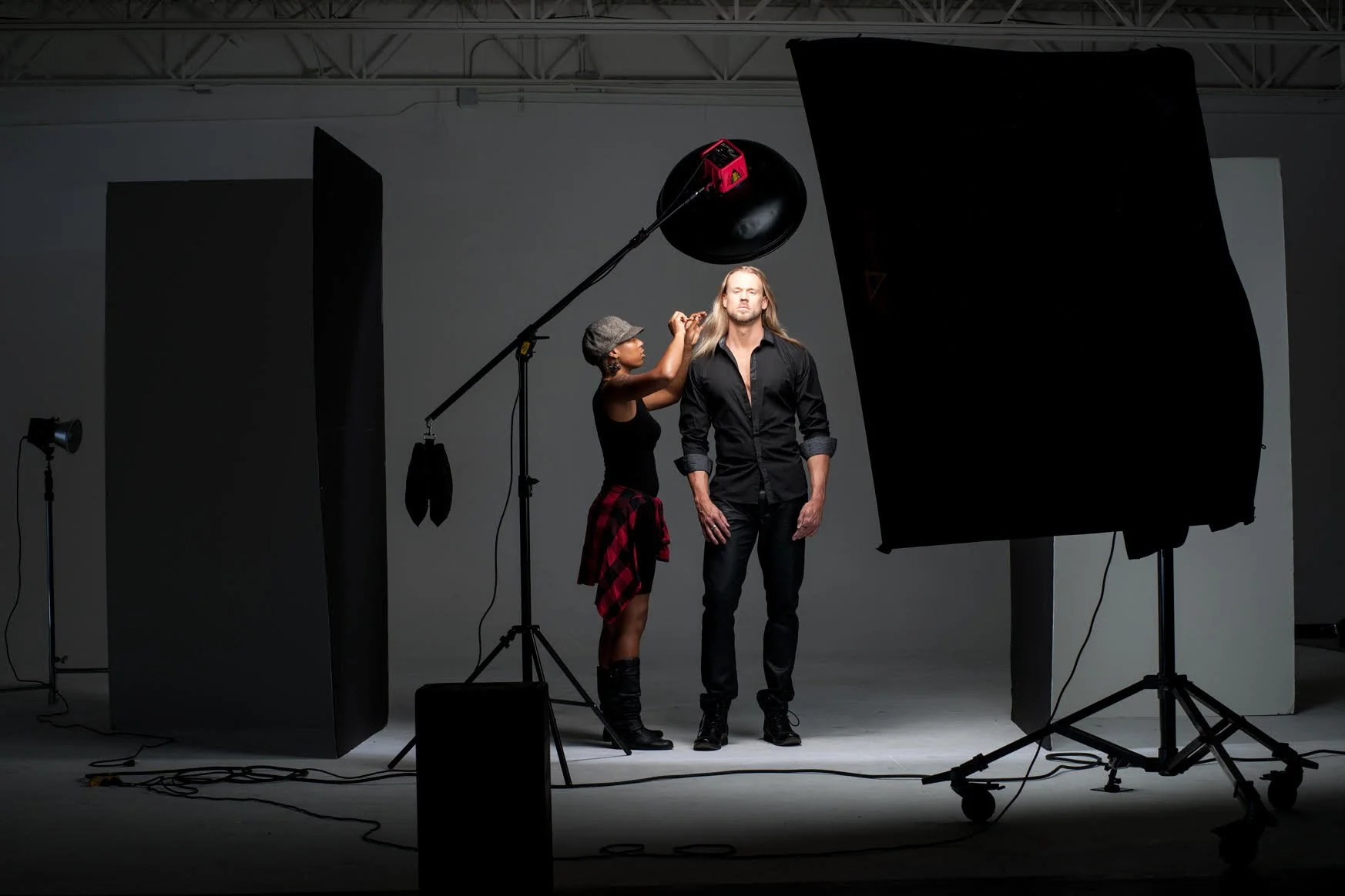 A professional photo shoot setup with a man and a woman posing on a gray background. The woman is touching the man's hair, both dressed in black, with studio lights and black reflectors around them.