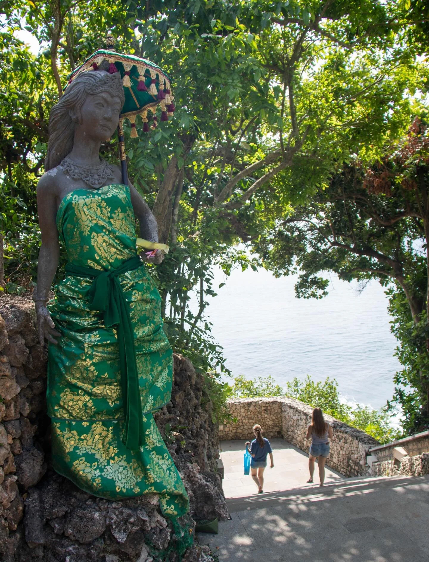 A quiet guardian on the path to the ocean &mdash; wrapped in green silk, holding offerings, and framed by sunlight through the trees. At AYANA, even a simple walk to the beach becomes a cultural experience to share as a family.

See our site for the 