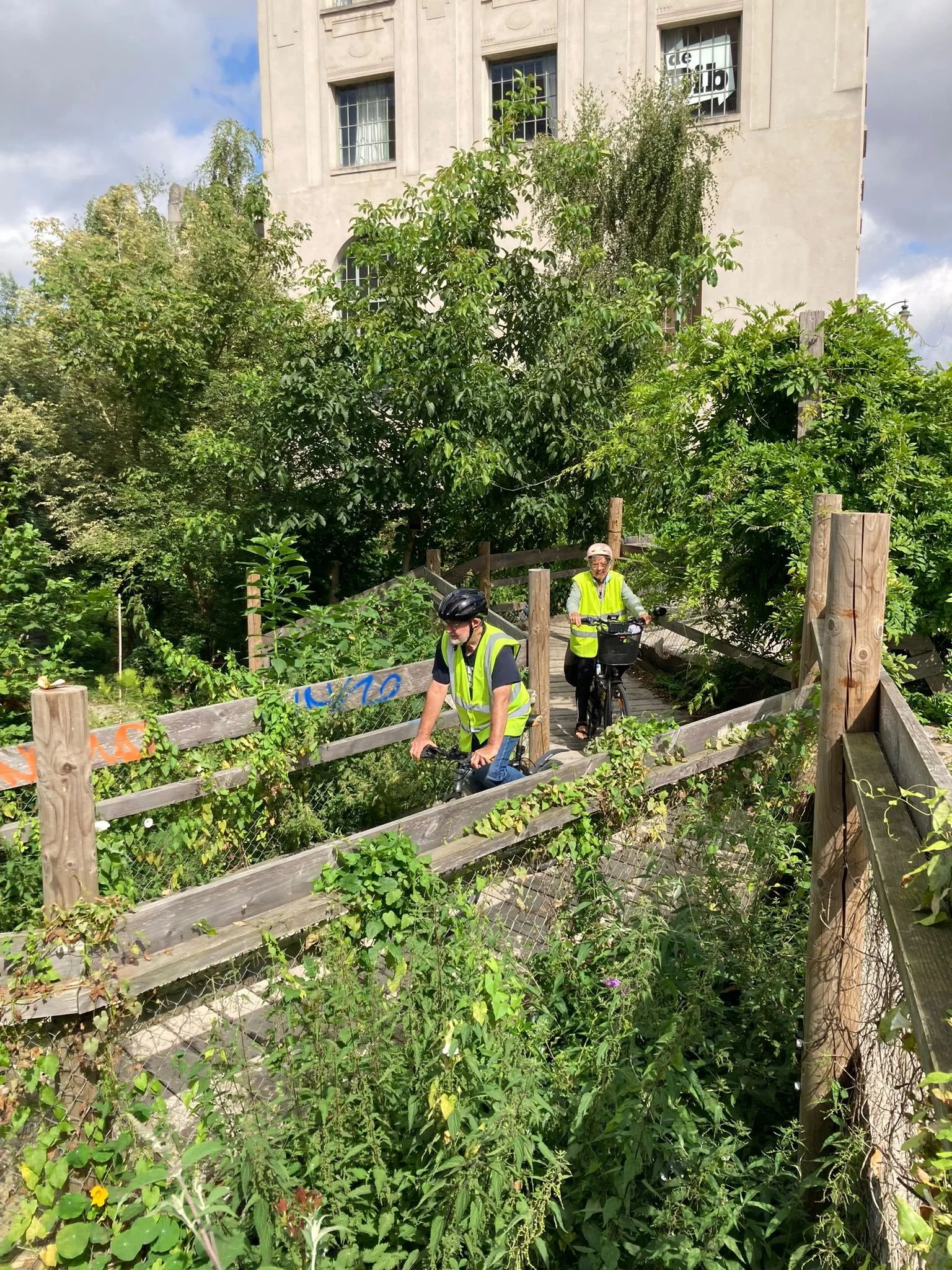 Twee mensen fietsen op een pad met houten en metalen hekken, omgeven door groen en bomen, met een gebouw op de achtergrond.