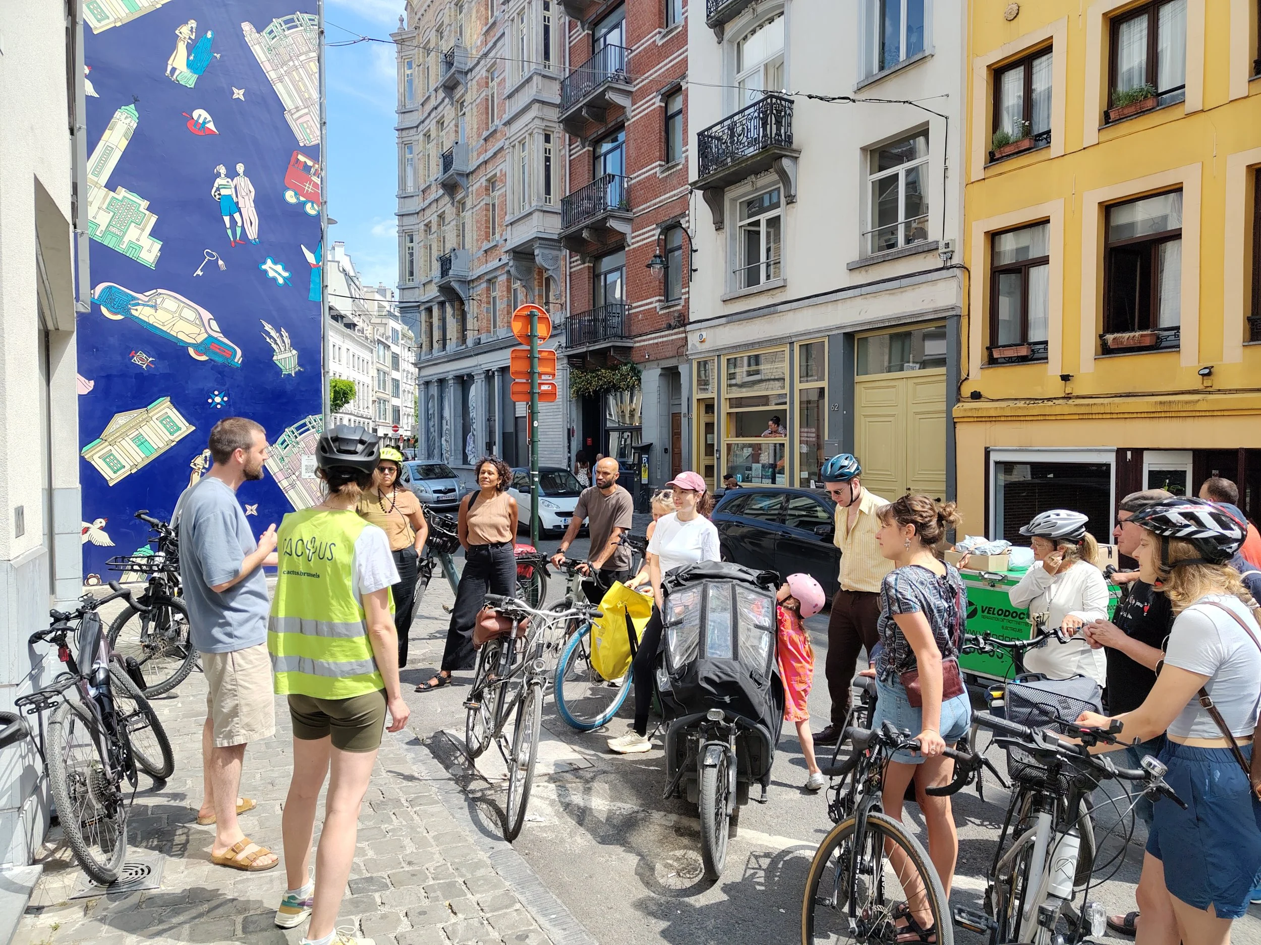 Mens en vrouwen op de straat met fietsen, omringd door historische gebouwen en een muurschildering, op een zonnige dag.