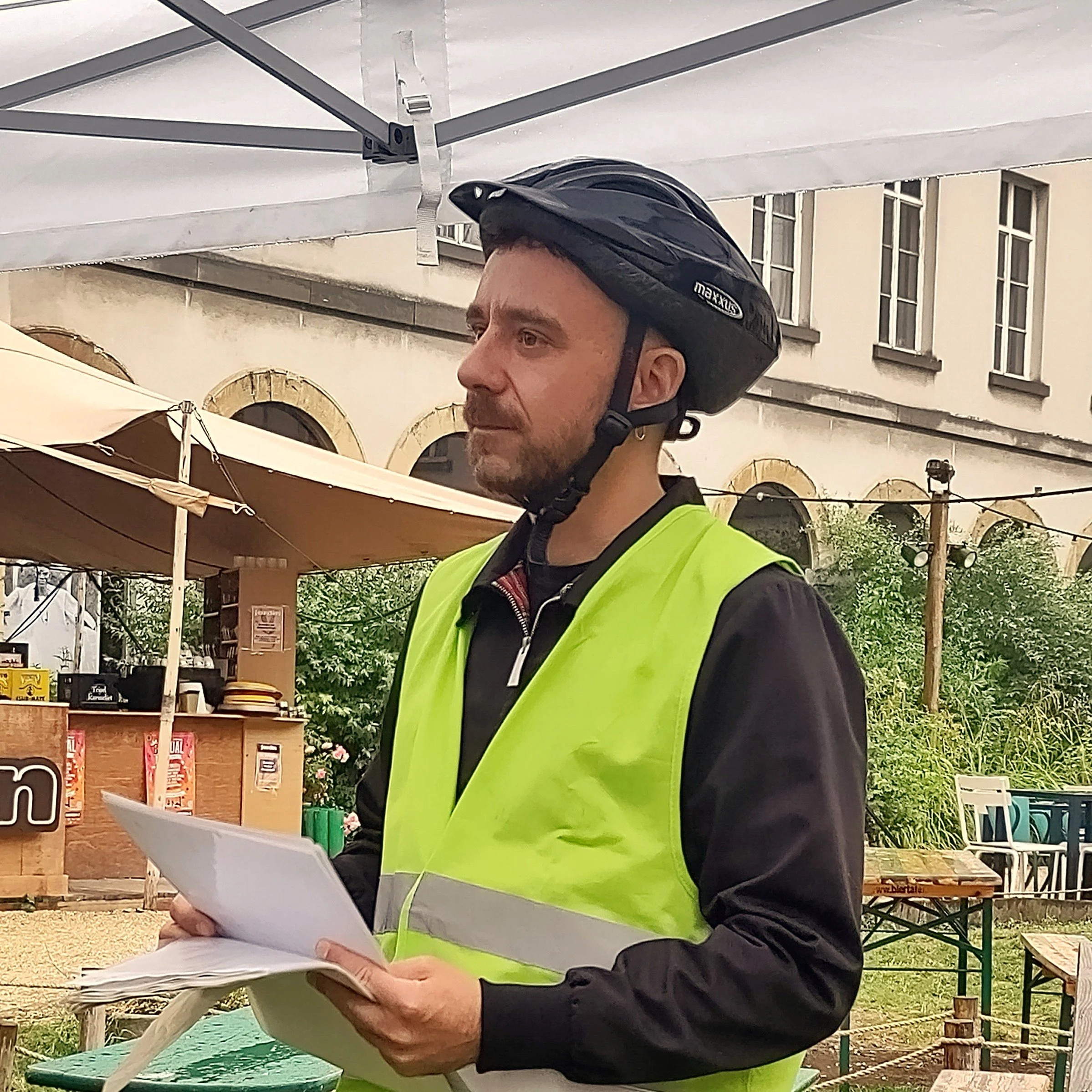 Man met een fietshelm en een gele veiligheidsvest die documenten vasthoudt, buiten onder een parasol.