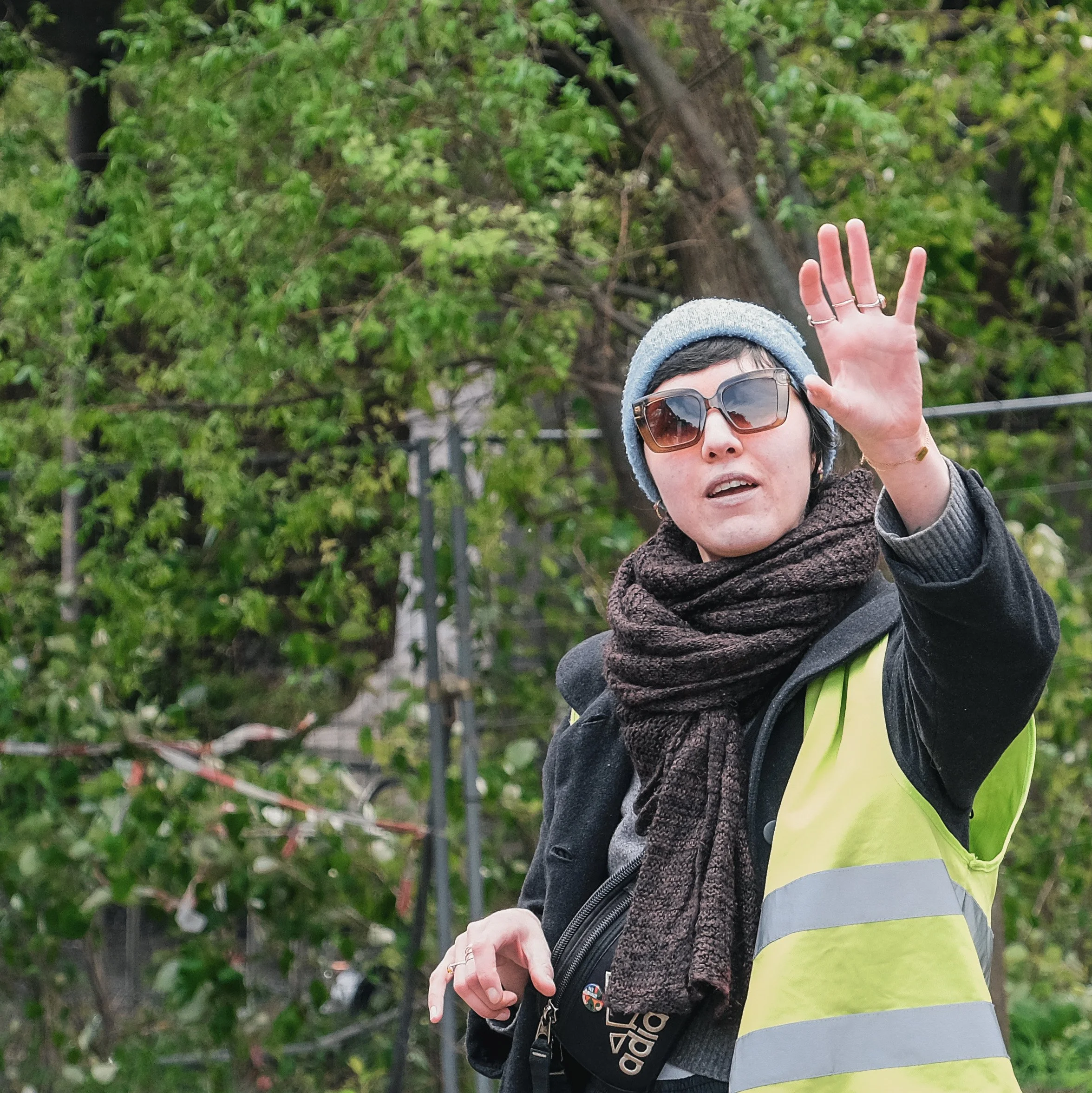 Vrouw met regenboogkleurige reflecterende veiligheidshes, donkere zonnebril, grijze beanie en een dikke sjaal zwaait met haar hand