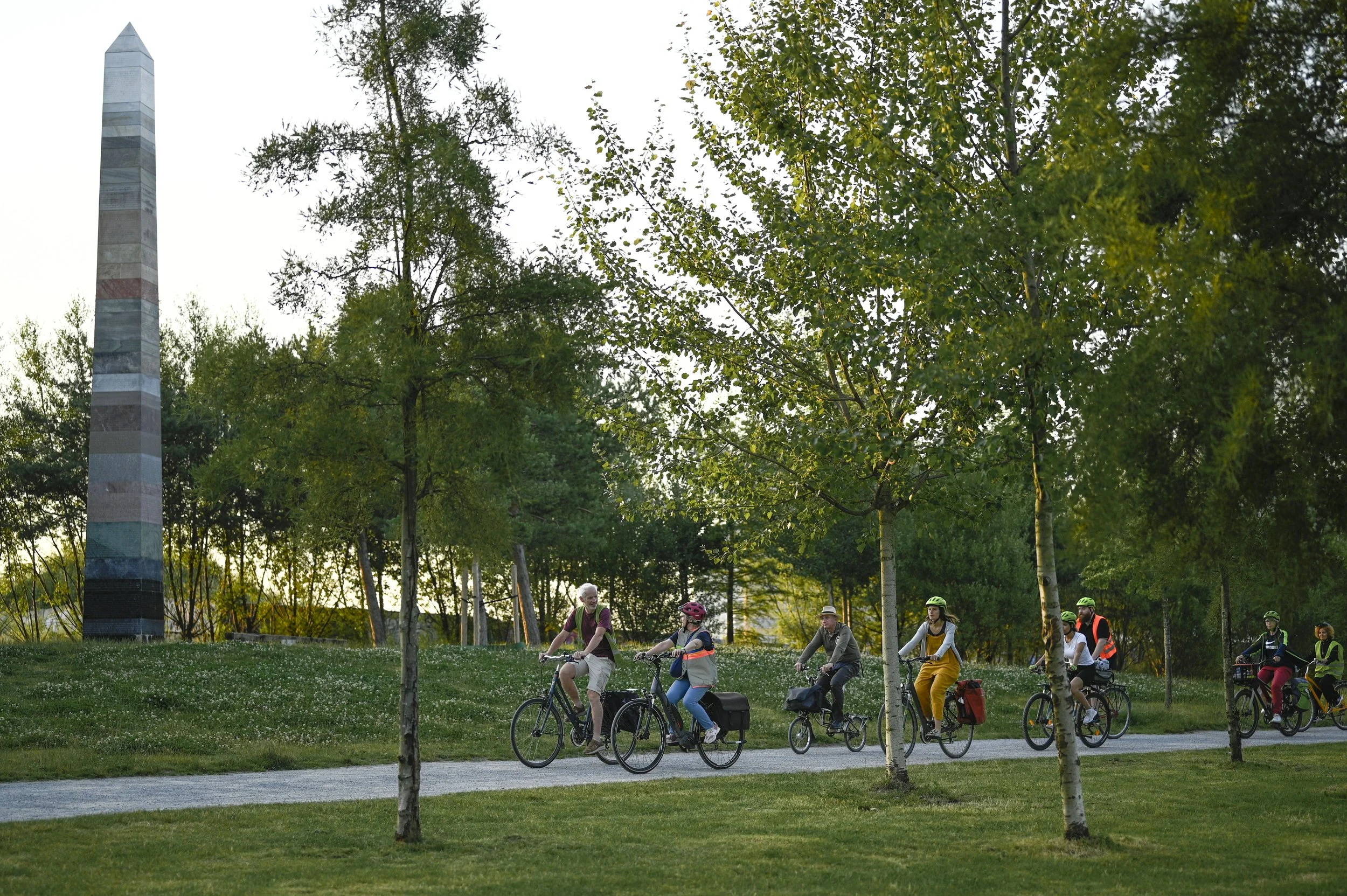 Een groep mensen fietst samen door een park met groene bomen en gras, terwijl de zon ondergaat.