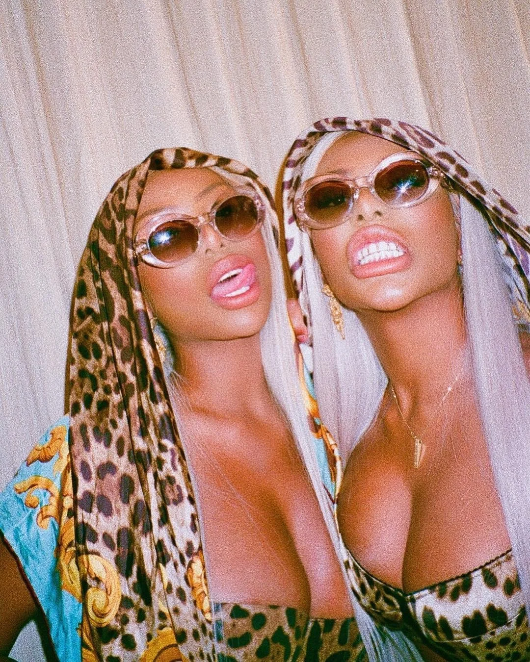 Two women with platinum blonde hair wearing leopard print scarves, sunglasses, and colorful outfits posing with facial expressions showing their grillz.
