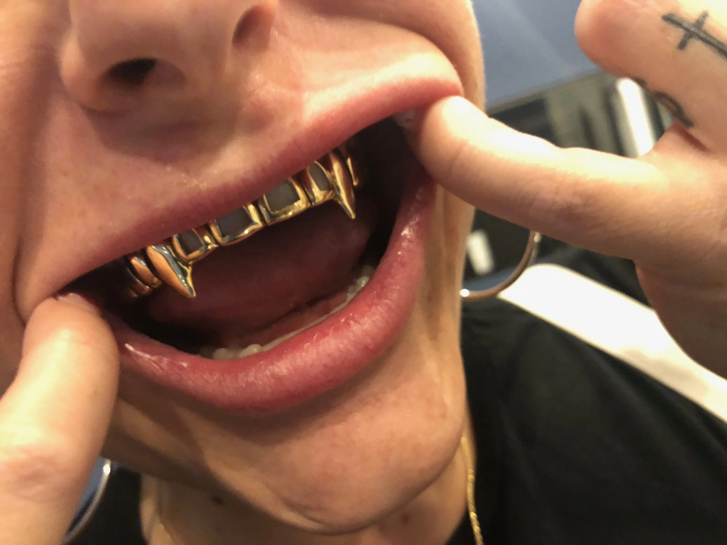 Close-up of a person with grillz with fangs on their canine teeth, opening their mouth and touching their face with fingers, showing the fangs on the grillz.