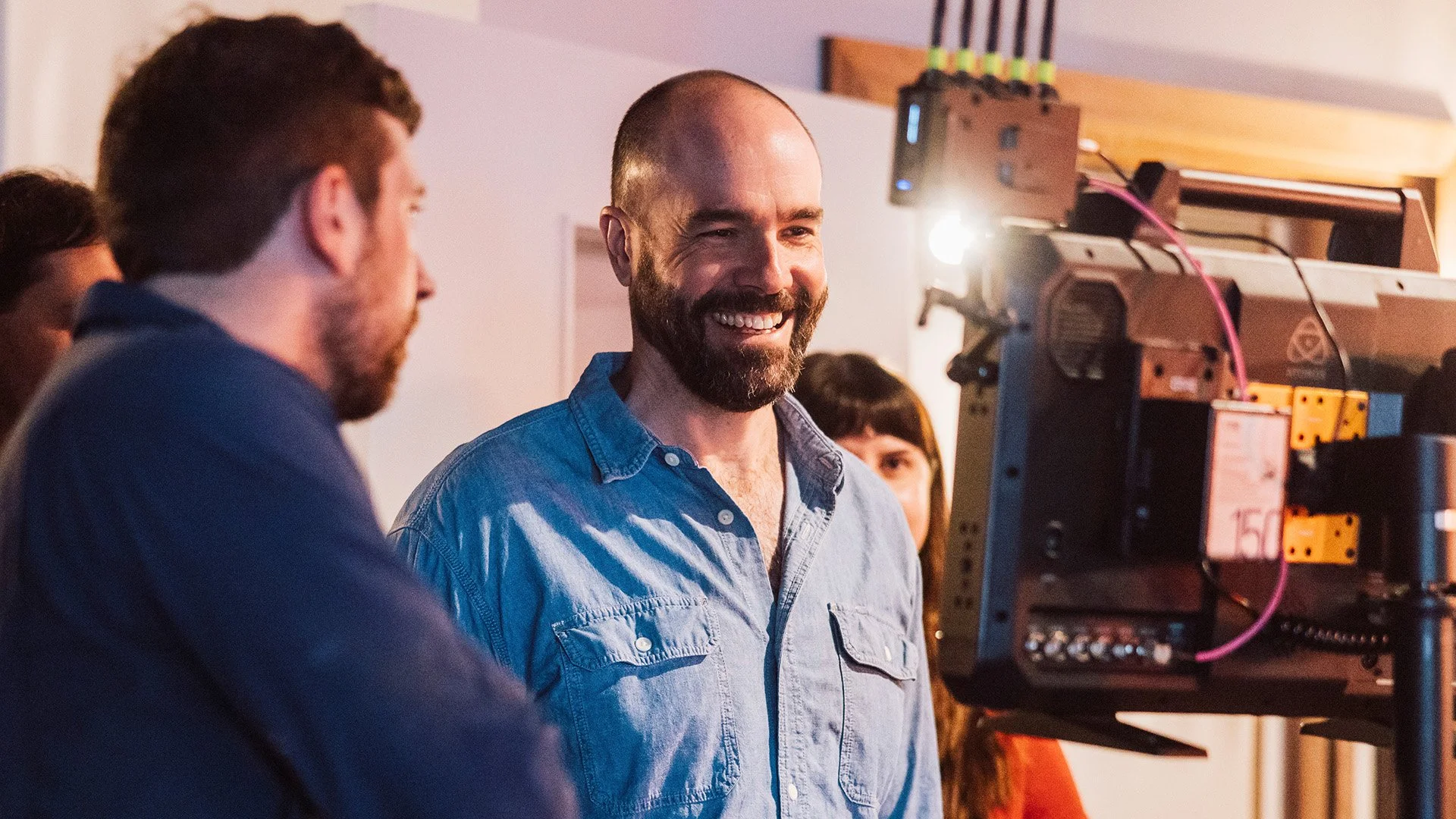 A group of people working together in a room, with a man, Michael O'Halloran, in a blue shirt smiling and looking at a piece of film equipment or camera.