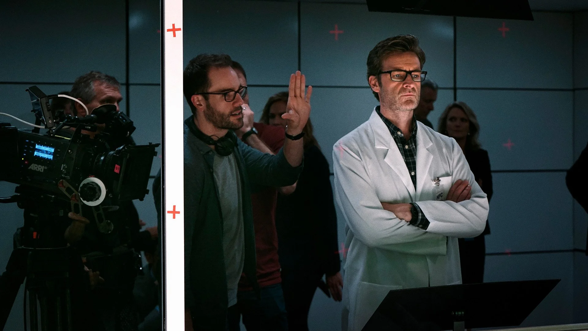 Behind the scenes of a film or TV production, a crew member, Michael O'Halloran, explains something to a doctor, who is wearing a white lab coat, in a studio or hospital setting.