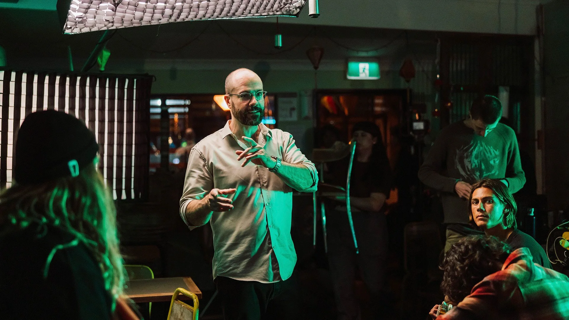 A man, Michael O'Halloran, with glasses, a beard, and a beige shirt speaking to a group in a dimly lit room with green and orange lights. Several people are seated and listening, with some looking at their phones or paying attention.