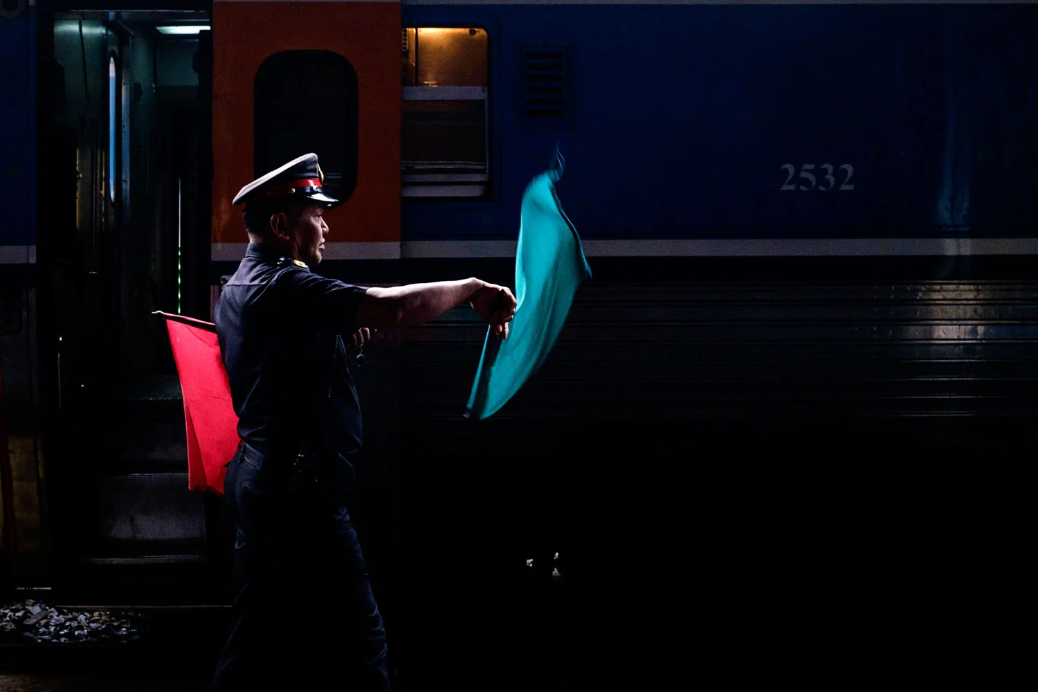 Near-black night frame on a railway platform: a guard in a peaked cap stands in profile inside a narrow band of light, one arm thrust out holding a turquoise flag caught mid-flutter and a red flag tucked under the other arm, against a deep blue carriage stencilled 2532 beside a slab of orange doorway