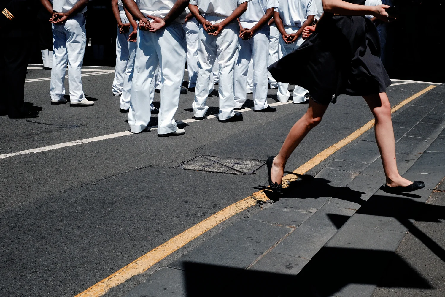 Harsh midday sun: a rank of sailors in white uniforms cropped off at the waist, hands clasped behind their backs, shadows pooled beneath them; at right a woman in a black skirt strides across a yellow kerb line, cropped at the shoulders, her shadow cutting a hard diagonal over the pavement