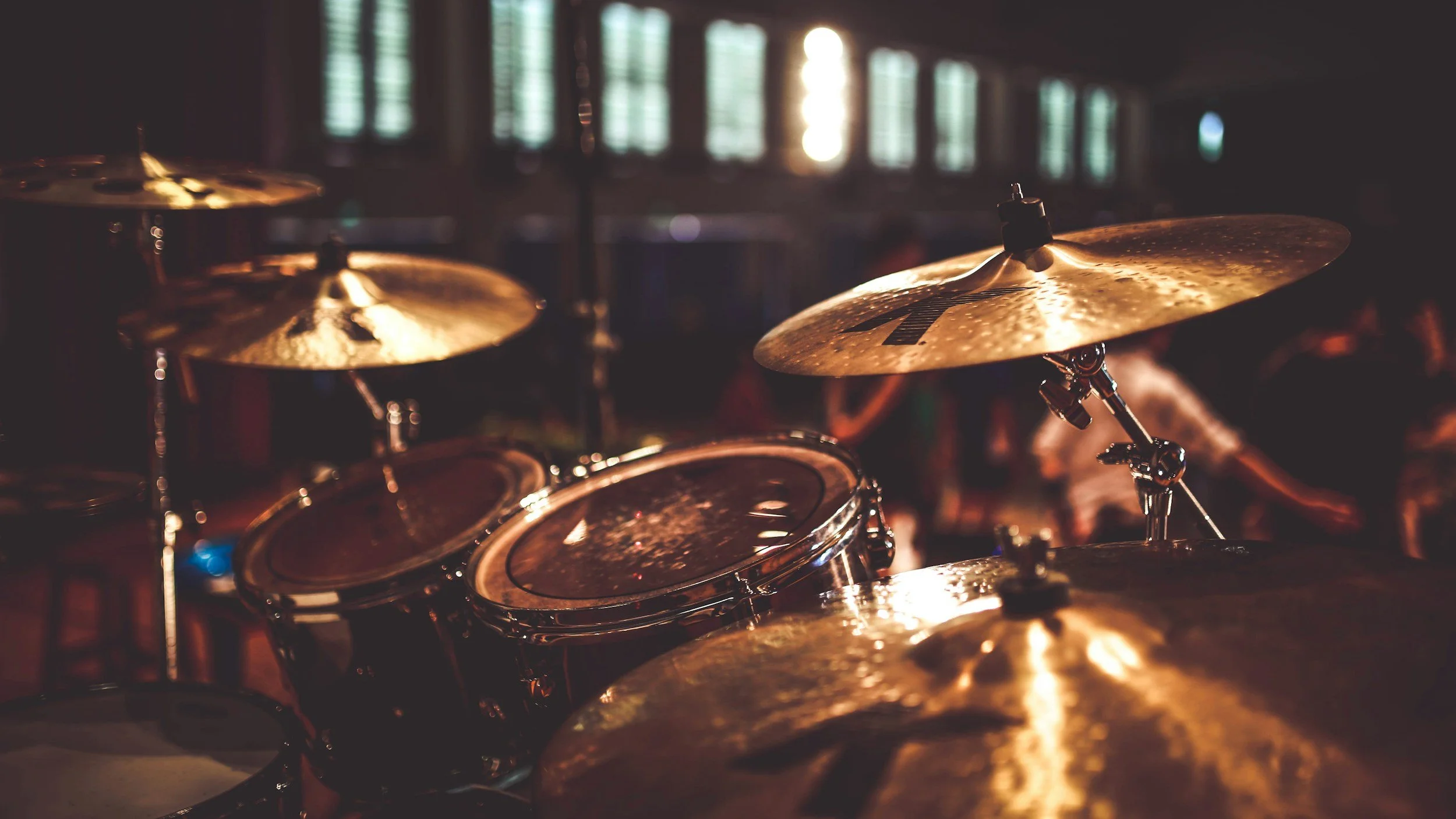 Close-up of a drum set in a dimly-lit room with sunlight filtering through windows in the background.