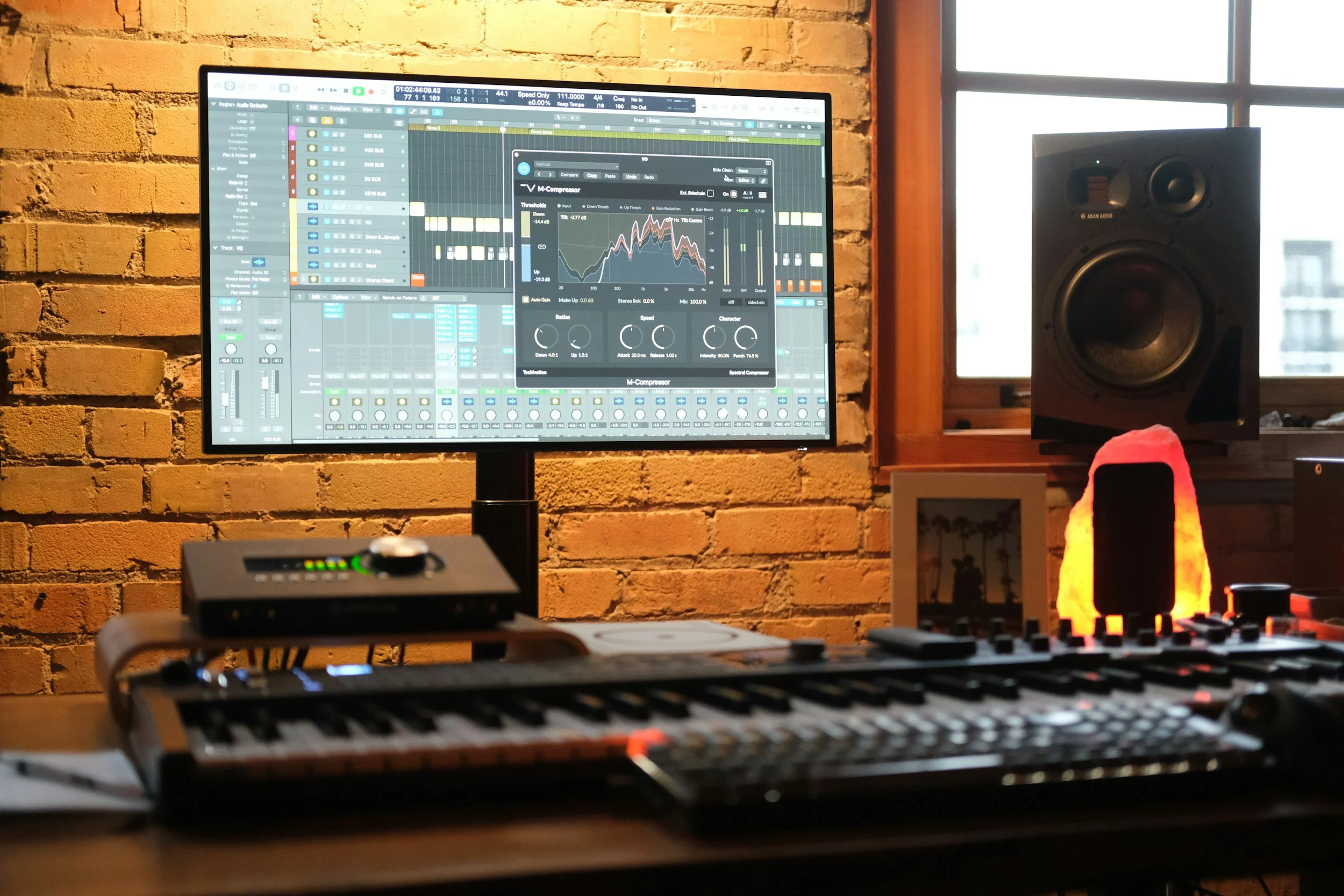 A computer monitor displaying music production software, with a speaker to the right, set against a brick wall and a window in the background.