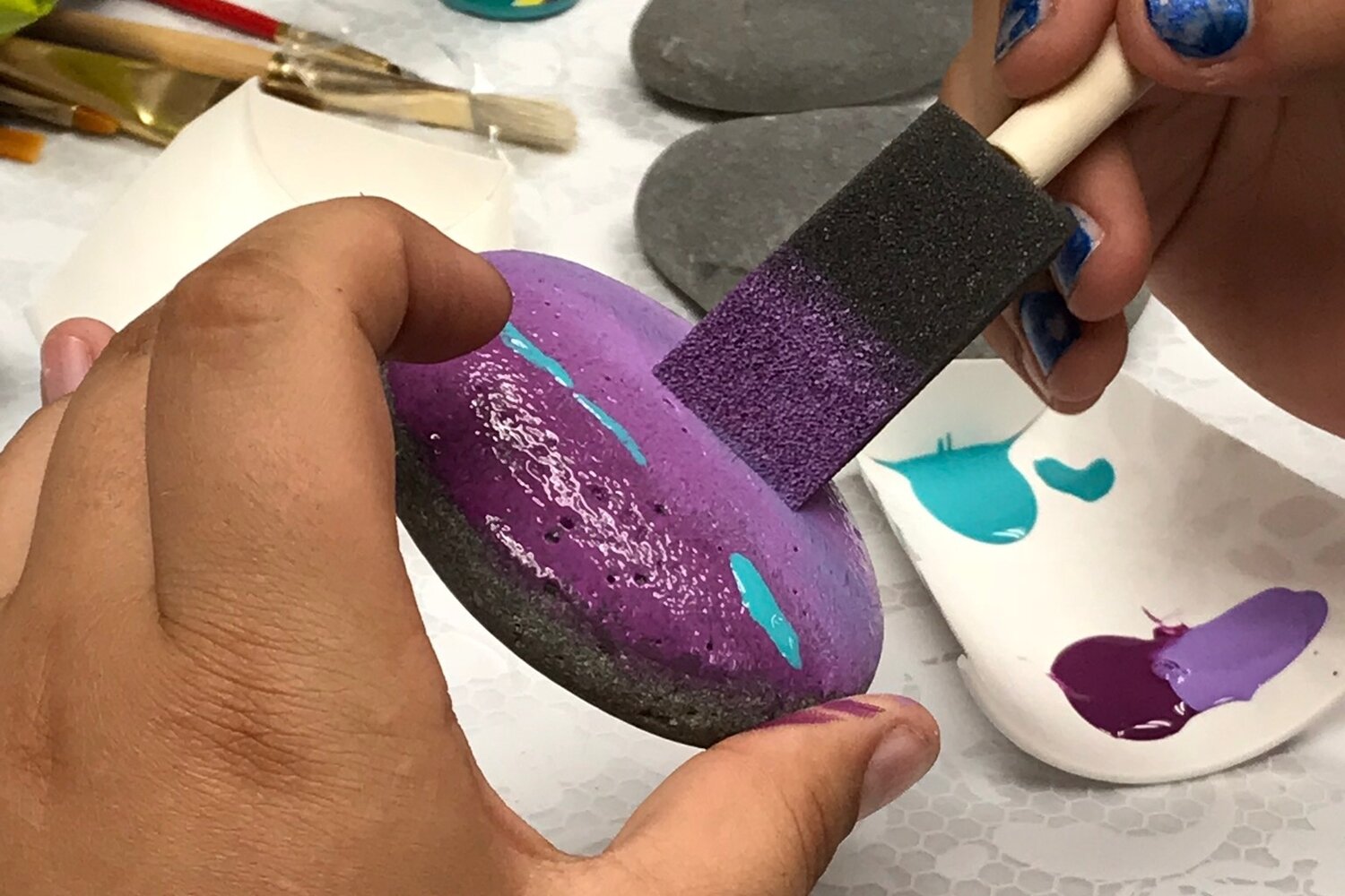  A rock is being painted purple with a blue stripe with a foam brush. The rock is held between the pointer finger and thumb of the painter. 