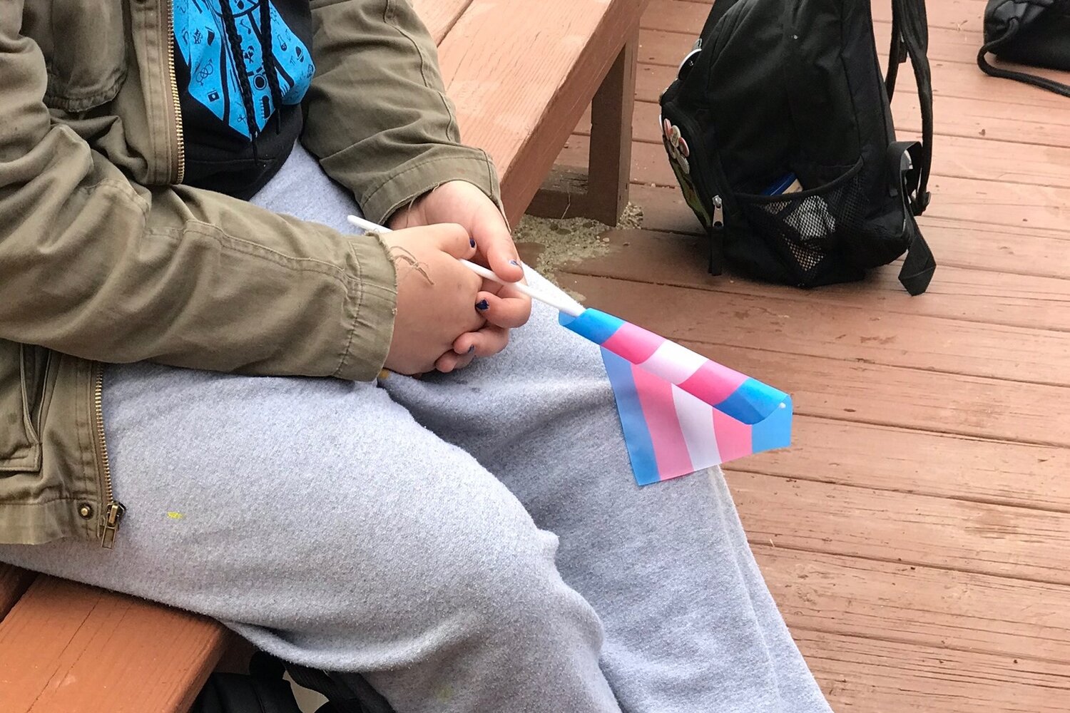  The bottom half of a teen wearing sweatpants and a green khaki sweater is seen holding the trans flag with both their hands. 