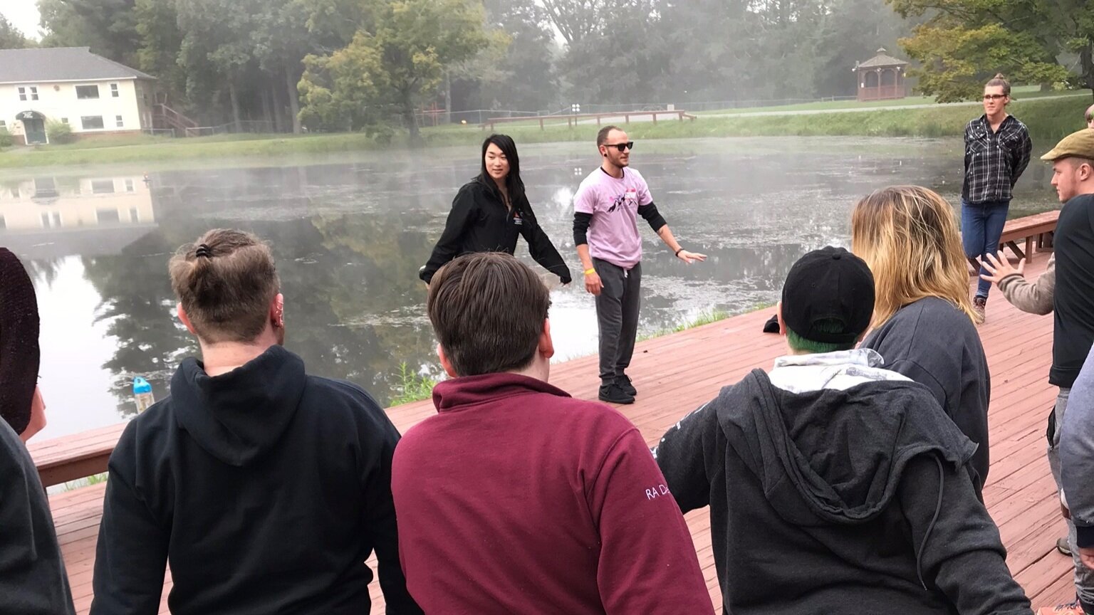  Two leaders stand on the edge of a dock on a lake. The backs of four individuals face the camera. 